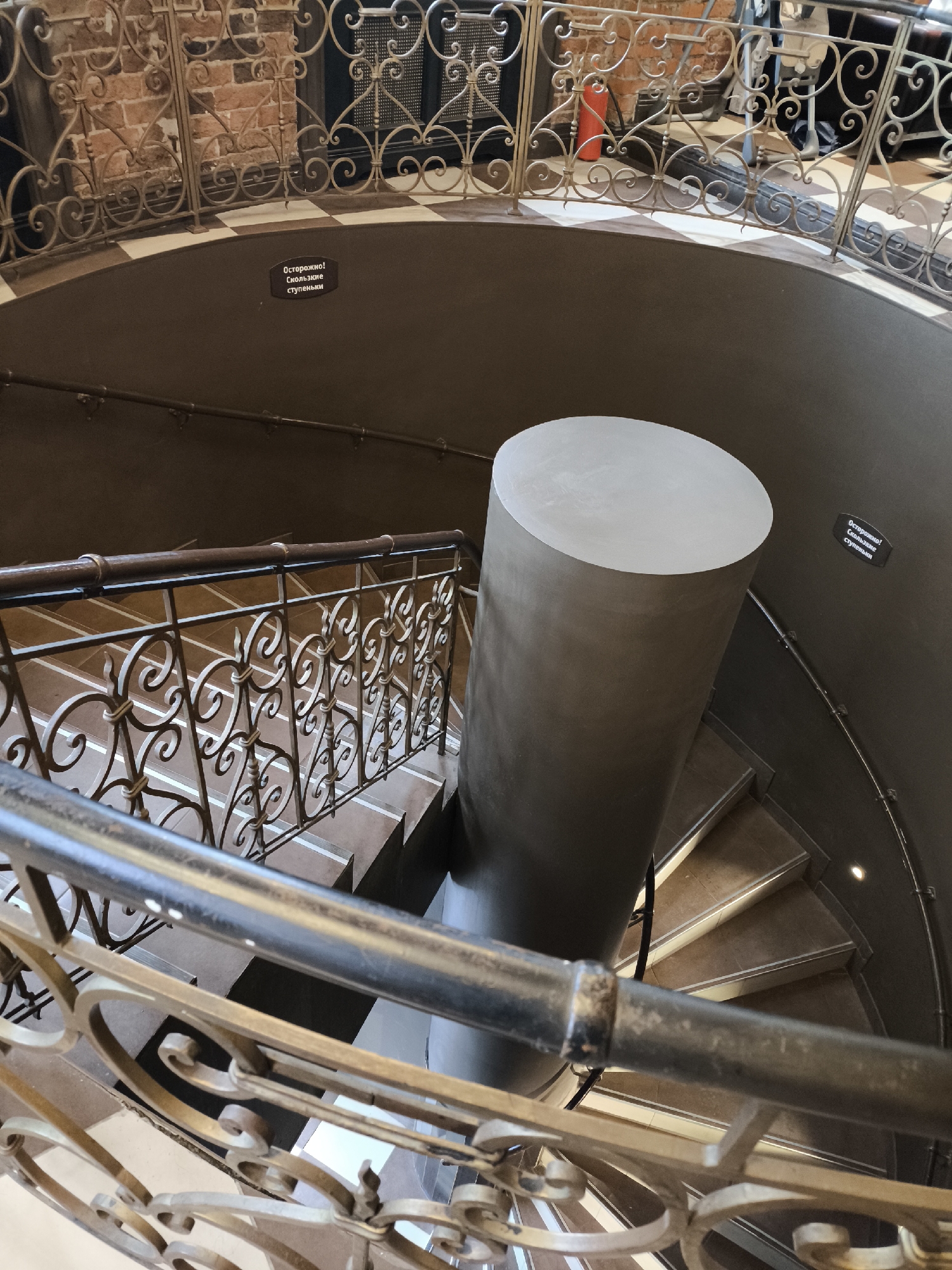 This image captures an interior view looking down into an elegant spiral staircase, likely within a grand or historic building in Moscow, Russia. The perspective is from an upper floor, overlooking the winding descent.
The central feature is a substantial, dark cylindrical column around which the staircase spirals downwards. The column's top surface, visible in the foreground, is flat and light grey. The steps themselves are made of a brown or beige material, possibly stone or wood, with contrasting lighter-colored edges or risers, and appear to be well-maintained.
The staircase is adorned with ornate wrought-iron railings featuring intricate scrollwork patterns, primarily in a dark, antique gold or bronze hue, topped with a smooth, dark handrail. These decorative railings line both the immediate foreground of the upper level and the visible descent of the staircase.
The walls of the circular stairwell are smooth and dark brown, providing a muted backdrop for the intricate ironwork. In the upper background, beyond the railings of the current level, a section of an exposed brick wall is visible, along with glimpses of a lighter-colored floor with a subtle checkered pattern, and additional decorative metalwork.
Two identical warning signs are visible on the dark wall of the stairwell. The text, written in Russian, reads: "Осторожно! Скользкие ступеньки," which translates to "Caution! Slippery steps." The overall lighting is bright, suggesting either daytime or well-lit interiors. No people are visible, and no specific activity is taking place other than the implied passage through the stairwell.