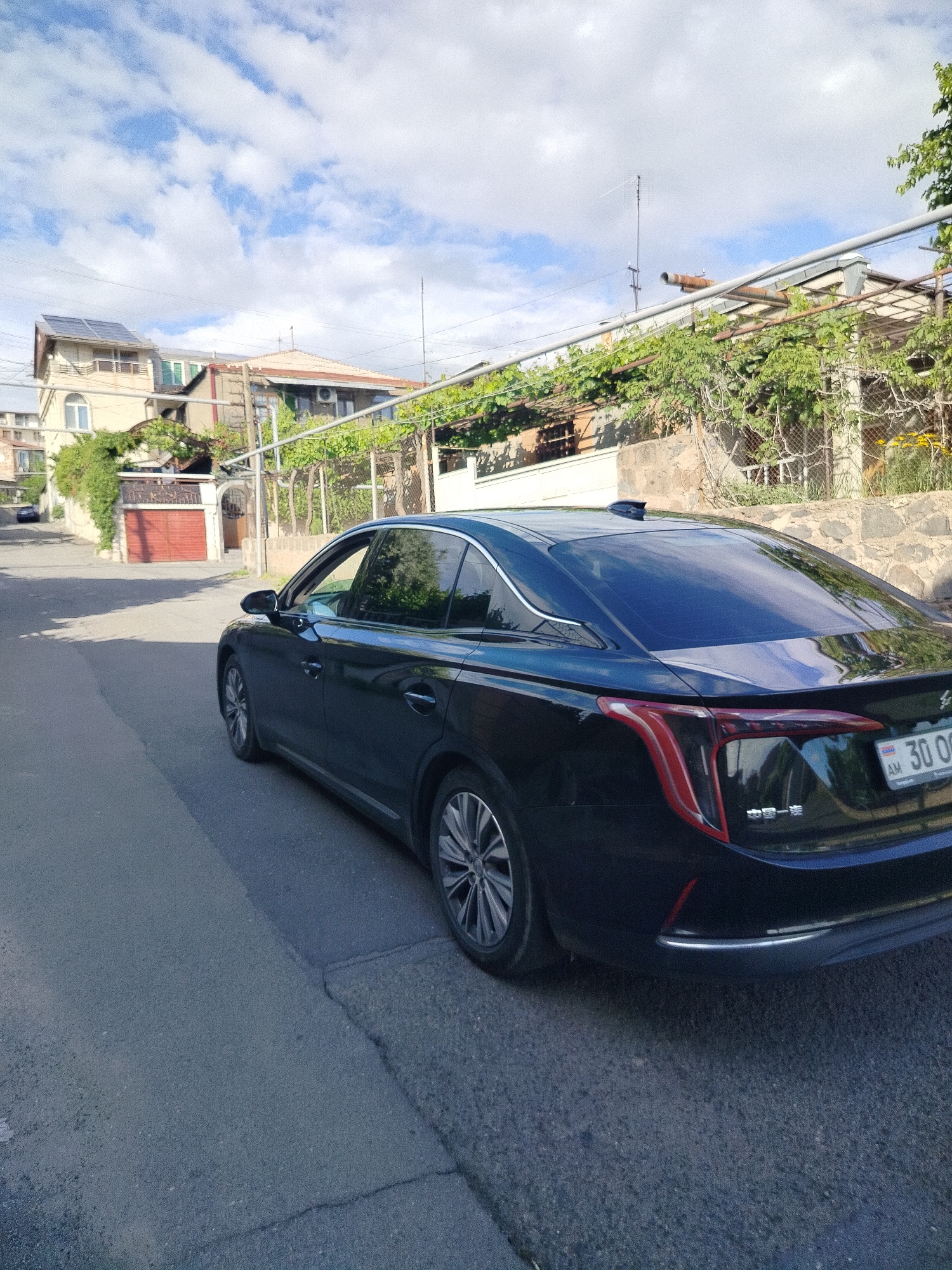The image shows a black car parked on a street in Yerevan, Armenia. The car, bearing an Armenian license plate ("30 OO"), is positioned on the right side of the frame, angled towards the viewer. To the left of the car is the street, and in the background, there are residential buildings, a stone wall covered in greenery, and a partially visible red garage door. Overhead are utility lines and a partly cloudy sky. The buildings appear to be older, and the environment is residential.