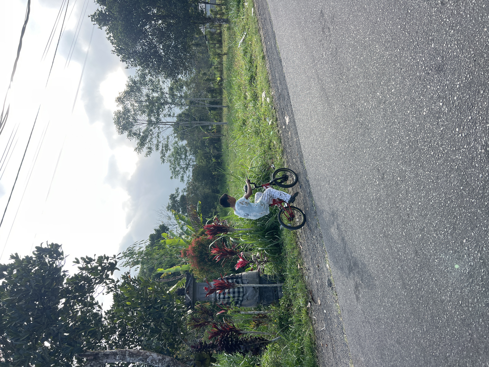 A young boy rides a red bicycle on a paved road in Rendang, Indonesia. The boy, wearing a light blue t-shirt and white pants with red accents, is positioned in the middle of the road, facing away from the camera and towards the right side of the frame. To his left, on the side of the road, is a stone monument adorned with checkered fabric. Lush green grass and vegetation surround the monument, with tall red-leaved plants and banana trees visible. In the background, more trees and foliage create a natural, rural landscape under a cloudy sky. The road itself is asphalt, with a visible shoulder of gravel and grass.