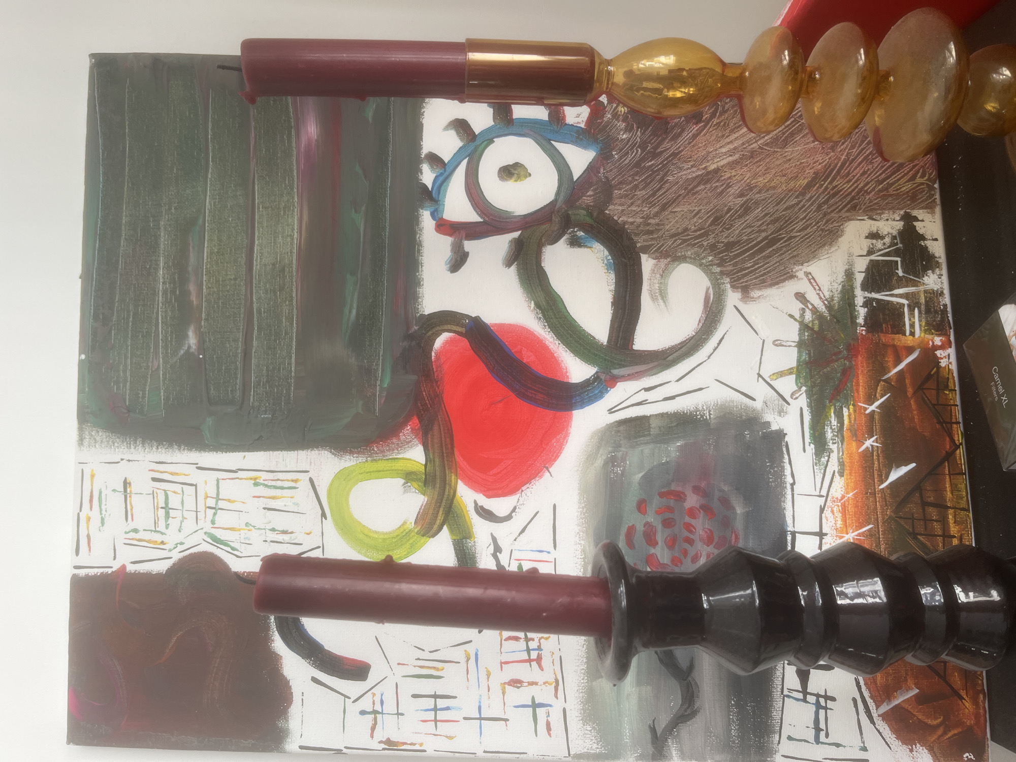 The image depicts a canvas with abstract painting, overlaid with two decorative candle holders and candles. The painting features a mix of dark green vertical strokes on the upper left, a central stylized eye with blue and brown outlines, a red circle, and swirling brushstrokes in green and brown. The lower portion of the painting contains a brown patch and a grid pattern in various colors. The canvas is partially obscured by the candle holders. The top candle holder is brown with a gold-colored, beaded ornament, while the bottom one is brown with a black glossy ornament. The candles inserted in the holders are both brown. The background appears to be a white surface, possibly a table or wall. A portion of what looks like a packaging box is visible in the upper right of the image. The location is Unknown, Unknown.