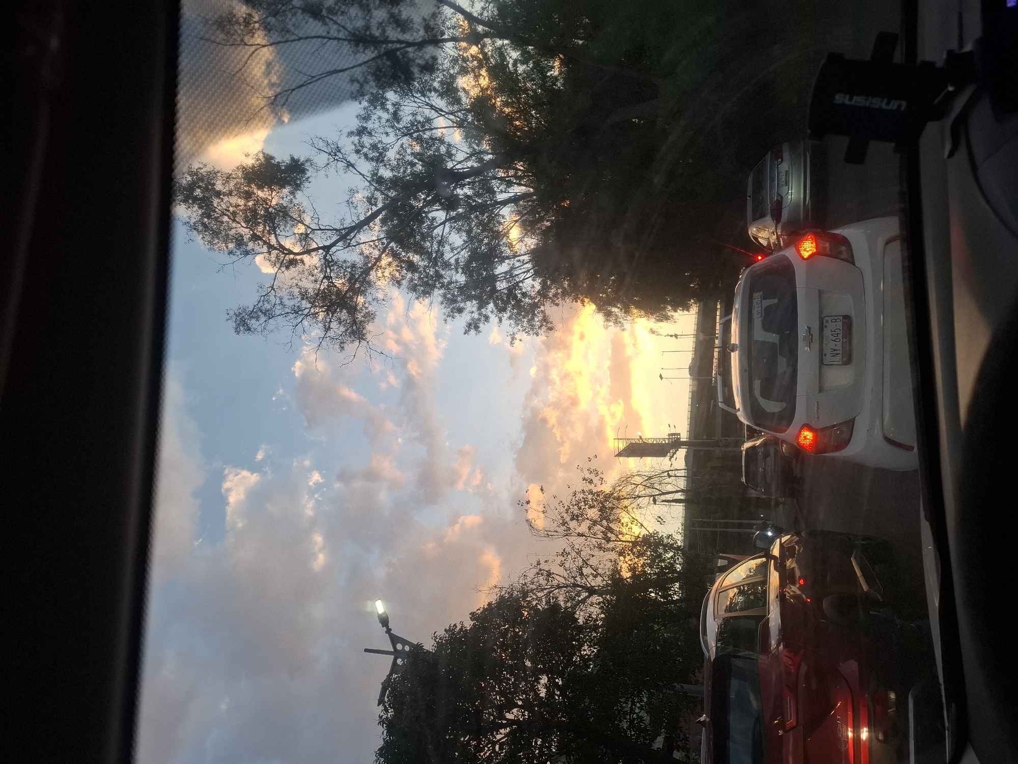 The image, taken from inside a vehicle, captures a traffic scene in Naucalpan de Juárez, Mexico, during a vibrant sunset. The sky is a mix of blue and orange-tinted clouds, framed by tree branches. Several vehicles are visible in a traffic queue, including a red car, a white car with license plate LNV-645-B, and a silver car. A black phone holder, branded "SUSISUN," is mounted on the dashboard. Streetlights are also visible along the roadside.