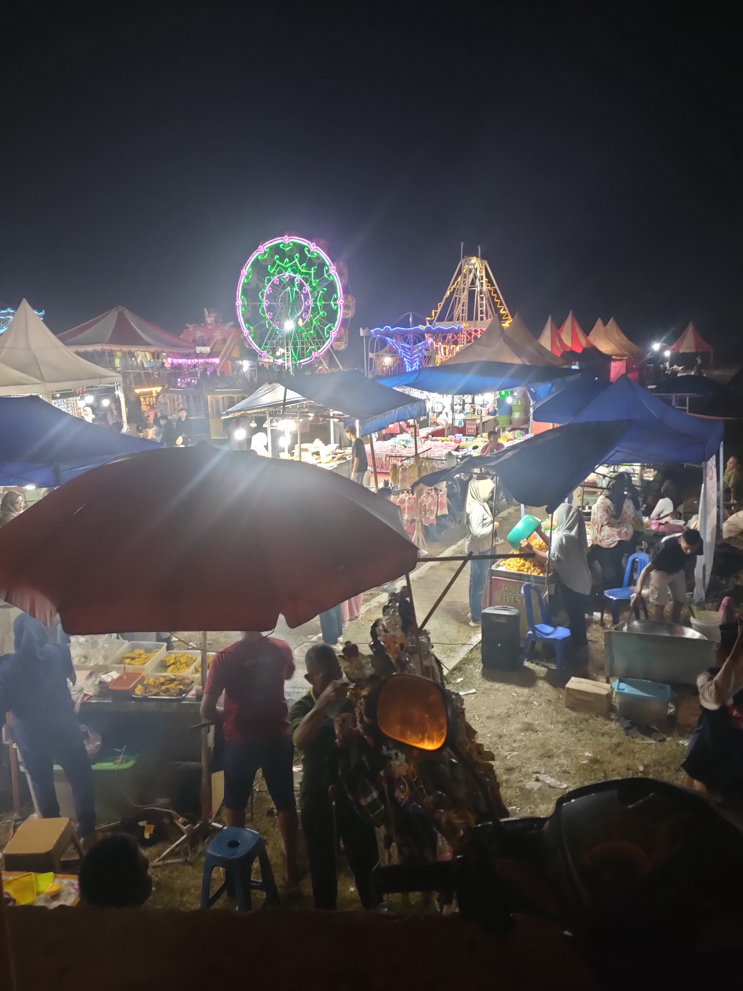 The image depicts a bustling night market scene in Subang, Indonesia, likely a temporary setup for a local fair or festival. Dominating the background is a brightly lit Ferris wheel, adorned with green and pink neon lights, suggesting a festive atmosphere. Adjacent to it, a tall, pyramid-shaped amusement ride, also illuminated with colorful lights, adds to the carnival ambiance. Various stalls and tents are set up throughout the market, covered with blue, white, and red canopies.
In the foreground and mid-ground, numerous people are engaged in various activities. Some are browsing or shopping at the stalls, while others appear to be vendors serving customers. One prominent stall features an array of fried food items displayed in containers. A person wearing a red shirt is standing behind this stall, seemingly attending to customers. Another stall has people gathered around, with one individual pouring something into a container, possibly preparing food or drinks. Blue plastic chairs are scattered around some of the stalls.
The ground of the market appears to be unpaved, with loose soil and scattered debris. A motorcycle is partially visible in the lower right corner of the image, its headlight illuminated. The overall lighting of the scene is a mix of the artificial lights from the amusement rides and stalls, creating a vibrant and somewhat hazy night market environment. The image captures a lively social gathering, with individuals enjoying the attractions and offerings of the market.