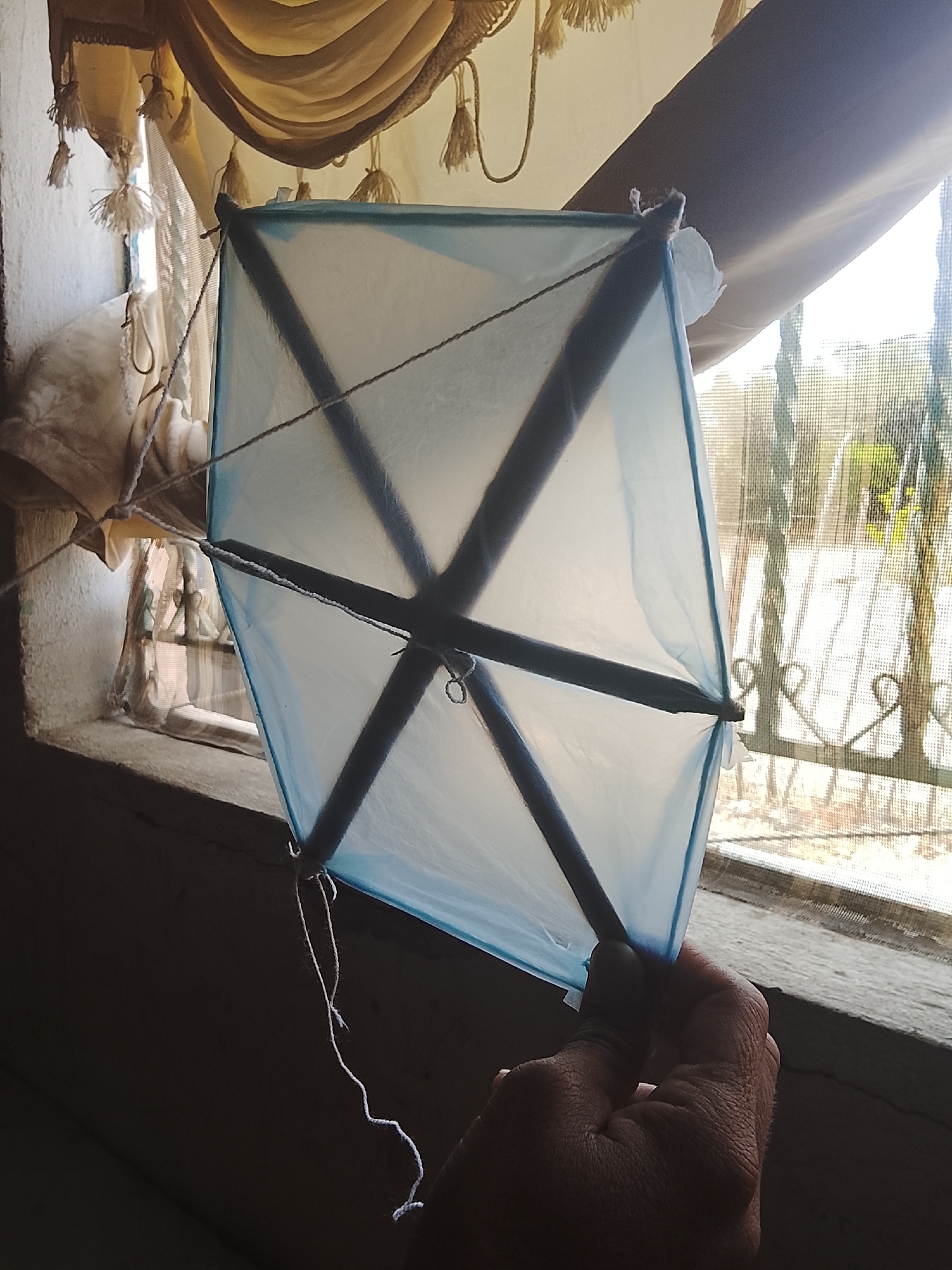 A hand holds a diamond-shaped kite with a light blue plastic covering and dark gray supports, indoors near a window. The window has a screen and overlooks an outdoor area with vegetation, possibly in Altagracia, Venezuela. Above the kite is a beige and white curtain with tassels. The window frame is rough and textured.