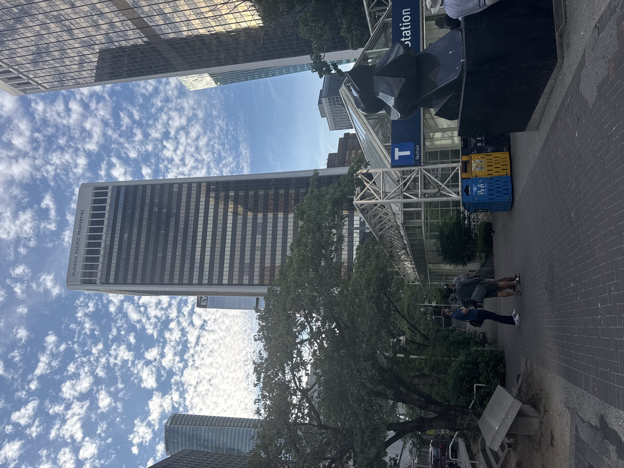 The image captures a street-level view of the Vancouver SkyTrain station at Granville Street and Georgia Street in downtown Vancouver, Canada, on a partly cloudy day. In the foreground, a black abstract sculpture sits on a granite base, with two people, one wearing a hat, standing near it. To the left, a paved walkway leads towards a park-like area with a stone marker and trees. Behind the sculpture and the walkway, a modern glass and metal structure houses the SkyTrain station entrance, indicated by a blue sign with a white "T" and the words "SkyTrain." Adjacent to the station entrance are three recycling bins: one blue, one yellow, and one black.
Dominating the background are several tall, glass-and-steel skyscrapers that characterize Vancouver's downtown core. The most prominent building features the "HUDSON PACIFIC PROPERTIES" logo at its apex. The sky is filled with scattered, wispy white clouds against a clear blue backdrop. The overall scene depicts a busy urban environment with a blend of modern architecture, public transit infrastructure, and elements of green space.