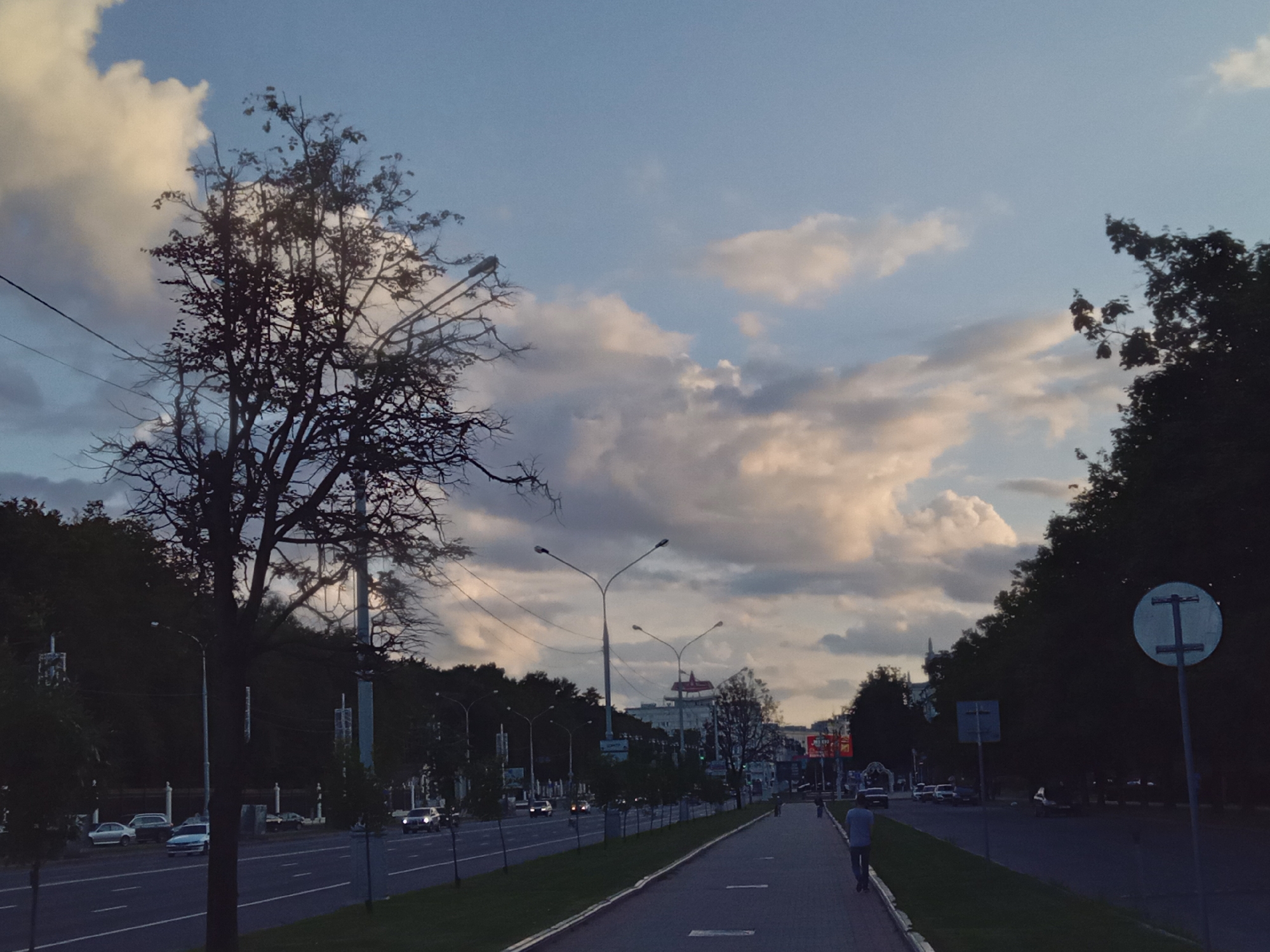 The image depicts a city street in Minsk, Belarus, during the daytime, with a mix of vehicles, pedestrians, and urban infrastructure under a cloudy sky. In the foreground, a paved pedestrian walkway runs alongside a multi-lane road. A lone male pedestrian is walking away from the camera on the walkway. On the road, several cars are visible, including a white SUV with its headlights on, moving away from the viewer. Trees line the sides of the street, with a large, bare-branched tree prominent on the left. Streetlights are visible along the road. In the background, buildings and taller trees are present, with a large red star visible atop one of the buildings, a common architectural feature in Minsk. A circular road sign with a white symbol on a blue background is positioned on the right side of the image. The overall atmosphere suggests an urban environment during a transitional part of the day, with dramatic clouds in the sky.