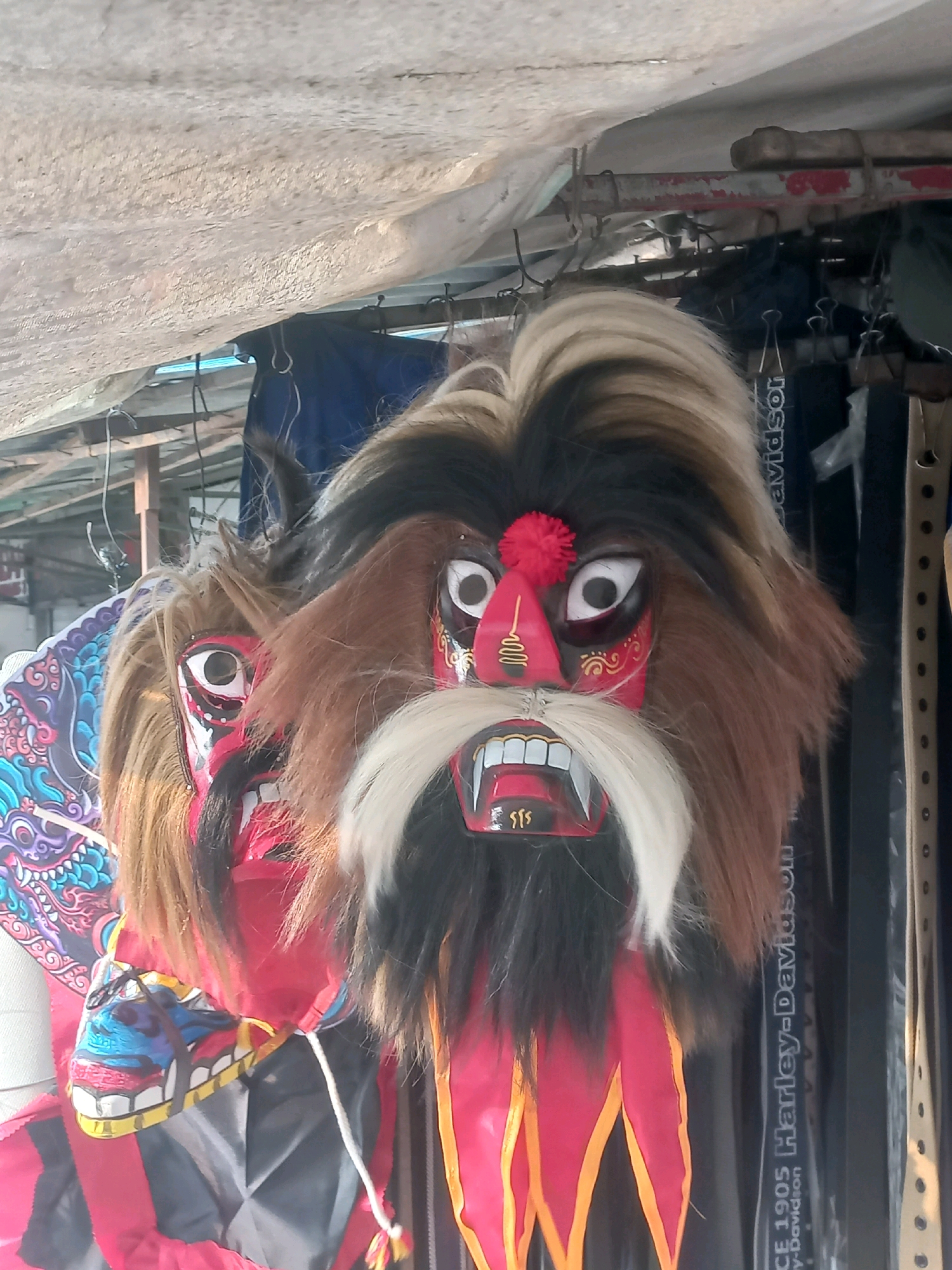The image shows two traditional Balinese Barong masks hanging from a structure, likely outdoors, with a weathered white tarp overhead. The masks are elaborate and colorful. The mask in the foreground has a prominent brown and black furry mane, striking white eyes with black pupils outlined in red, and a bright red face with black accents and yellow markings. Its mouth is open, revealing sharp white teeth and a red interior. A white beard and mustache frame the mouth. A small red pom-pom sits on its forehead. Behind it, another mask is partially visible, also with large eyes and red facial markings, and a blue, black, and white decorated headdress or headpiece. The masks are likely part of a display or performance costume. Behind the masks, various dark objects and straps are visible, including one strap with the text "Harley-Davidson" printed vertically. The overall impression is of a vibrant and somewhat chaotic setting, possibly a market or performance venue in Baturetno, Indonesia.