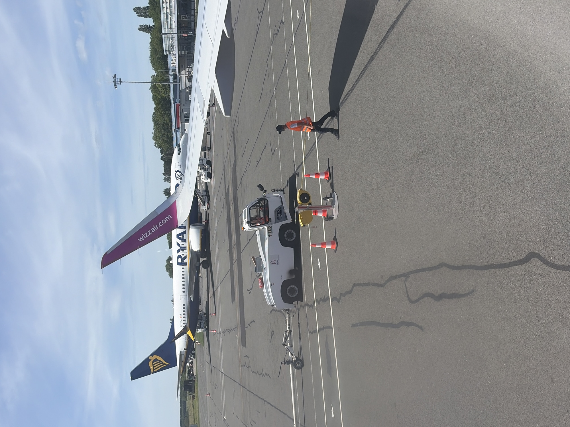 The image captures an airport scene in Tillé, France, featuring two commercial airplanes, one a Ryanair and the other a Wizzair, parked on the tarmac. A tow truck is positioned near the Ryanair plane, suggesting an imminent or ongoing aircraft handling procedure. An individual wearing a high-visibility vest walks near the tow truck and the aircraft, seemingly involved in ground operations. The background shows airport infrastructure, trees, and a blue sky with white clouds. The tarmac is marked with white lines. Traffic cones are also visible, likely placed for safety or guidance purposes.