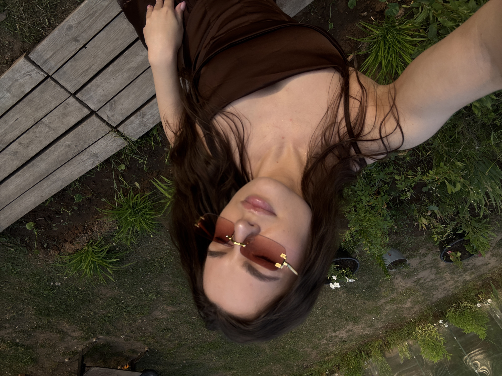 A woman is taking a selfie outdoors, sitting on wooden planks. She has long, dark brown hair and is wearing a brown dress and rimless sunglasses with pink lenses. The background shows a grassy area with plants and a greenhouse. The location is in Rusalevka, Russia.
