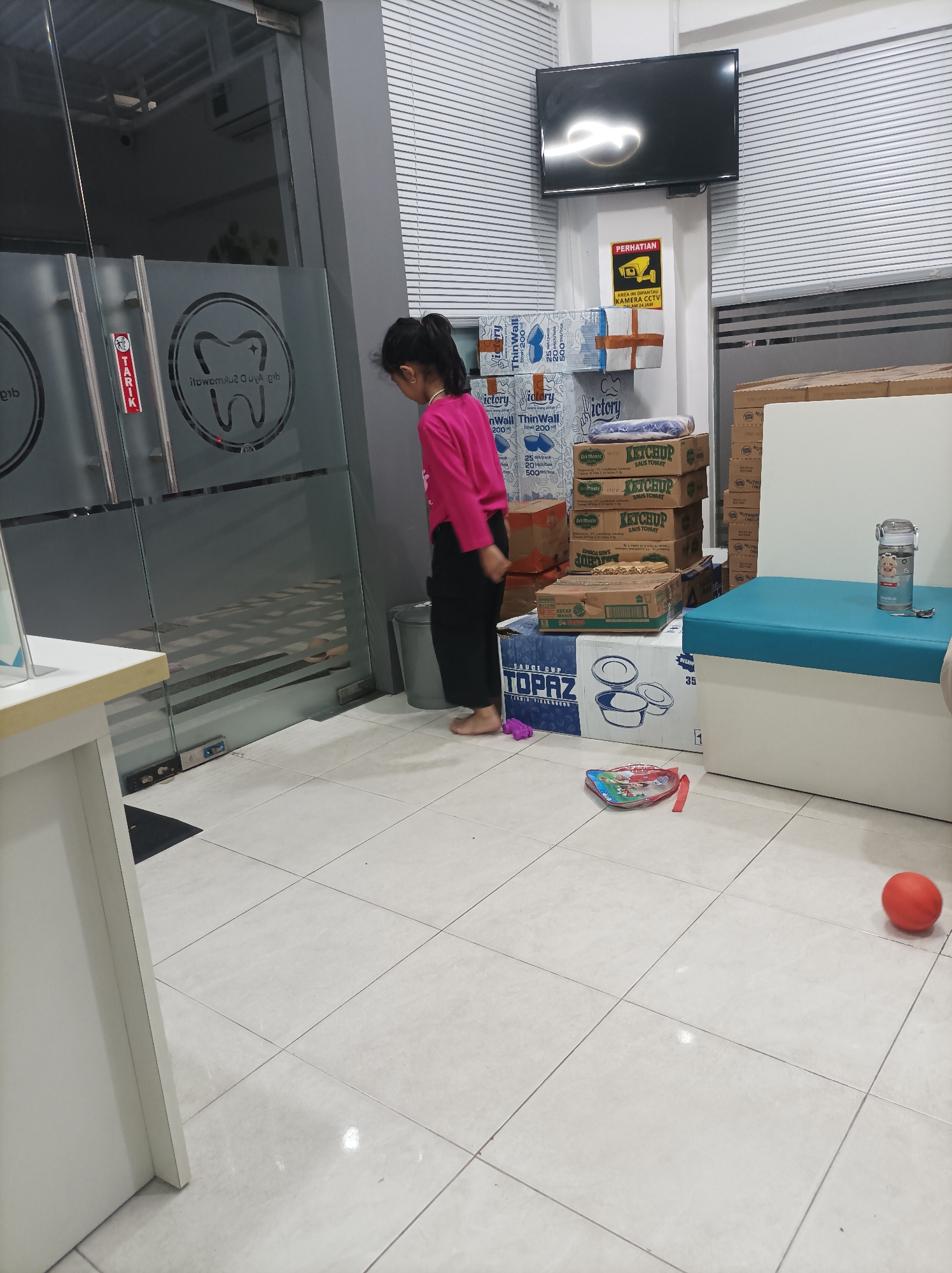 A young girl in a pink shirt and black pants stands in a brightly lit, tiled room, possibly a waiting area at a dental clinic, with a sign depicting a tooth logo on the glass door. She faces away from the camera, looking towards the door. Several stacks of boxes are visible, some labeled "Victory ThinWall" and "Ketchup" with the brand "Del Monte". A security camera sign is mounted on the wall above the boxes. A television is mounted high on the wall, and a child's toy lies on the floor near a bright red ball. The room appears to be indoors, with horizontal blinds covering a window or another wall. The floor is made of light-colored square tiles. The overall impression is of a clean, orderly space, likely a commercial establishment.