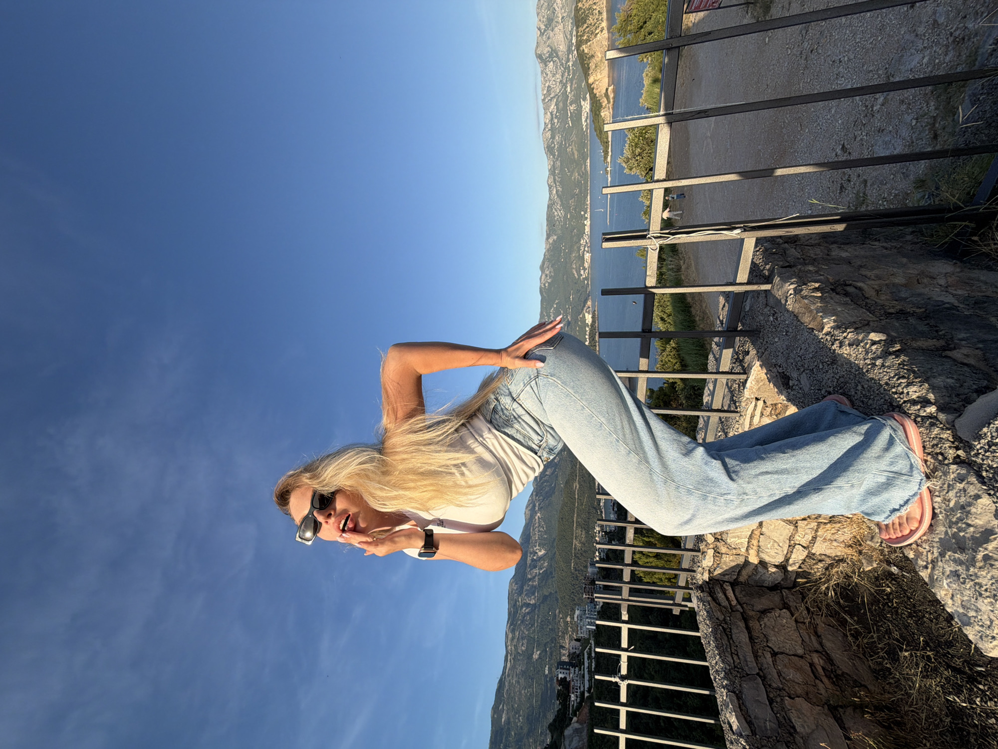 A blonde woman is standing on a stone ledge overlooking the city of Budva, Montenegro. She is wearing sunglasses, a white t-shirt, and blue jeans. She is making a kissing gesture towards the camera. In the background, there are mountains, the Adriatic Sea, and buildings. The sky is clear and blue.