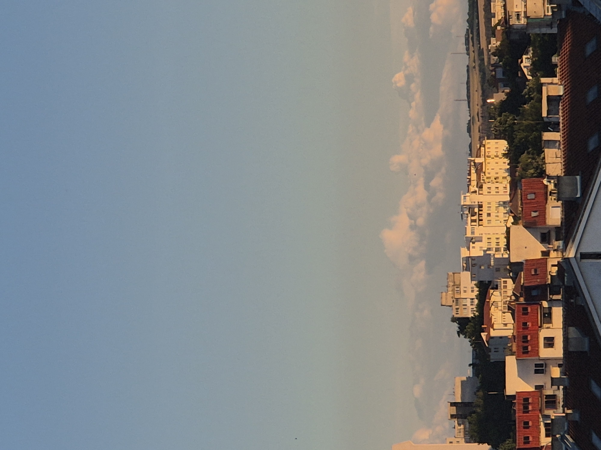 The image depicts a cityscape bathed in the warm light of a setting or rising sun. The sky is a clear, soft blue, transitioning to a lighter hue near the horizon. Scattered, fluffy clouds are illuminated by the sun, creating soft pink and orange tones. In the foreground and middle ground, a dense collection of buildings is visible. These structures are of varying heights and architectural styles, many featuring red-tiled roofs. Some buildings are multi-story apartment blocks with visible windows, while others appear to be smaller residential or commercial structures. Green trees are interspersed among the buildings, suggesting a developed urban area with some green spaces. In the distance, a rolling landscape of fields can be seen, punctuated by what appear to be communication towers or structures. The overall impression is one of an urban panorama observed from an elevated viewpoint, likely a rooftop or a higher floor of a building, during the golden hour.