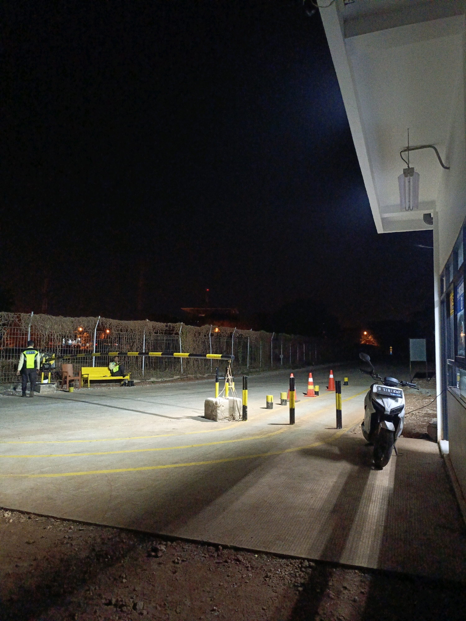 The image depicts a nighttime scene at a security checkpoint, likely in the vicinity of a facility in Jenu, Indonesia, potentially near a port or industrial area given the presence of a barrier and security personnel.
In the foreground, a paved road with yellow lane markings is visible, leading towards a closed barrier. To the left, a security guard in a yellow reflective vest stands near a gate control panel. Further back, another security guard sits on a yellow bench next to the barrier. The barrier itself is a black and yellow striped pole, lowered to block passage. Orange traffic cones are placed on the road, along with bollards.
To the right of the road, a white motorcycle is parked. The motorcycle has a license plate that reads "B 5181 KWF." Beyond the road, a wire fence topped with barbed wire runs along the perimeter. In the background, faint lights suggest the presence of distant buildings or infrastructure. The scene is illuminated by artificial lighting, casting shadows across the ground. To the far right, the corner of a building with a white overhang and a light fixture is visible. The overall atmosphere is one of security and control.