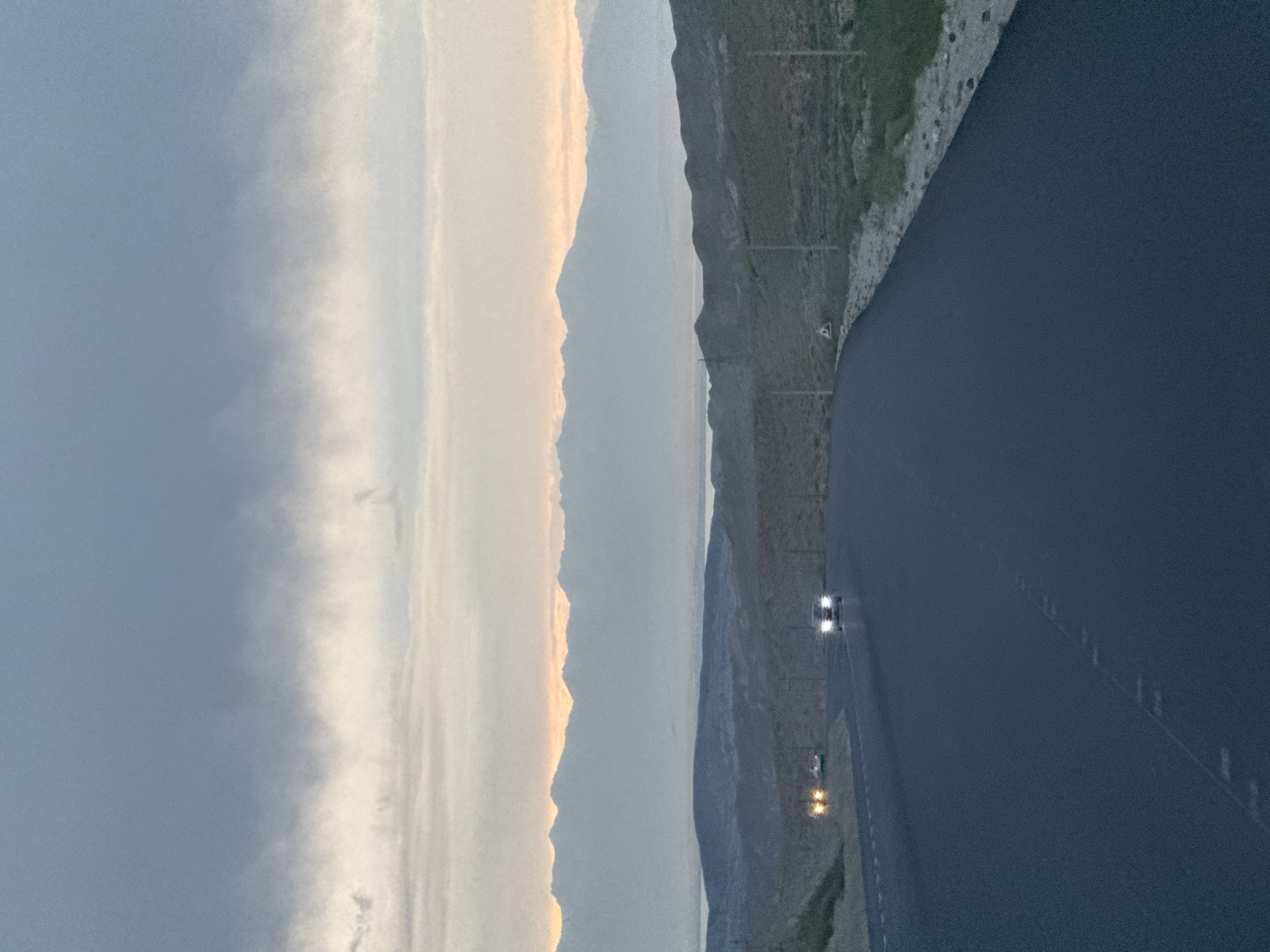 An asphalt road curves through a landscape of rolling hills under a cloudy sky with hints of orange from a setting or rising sun. In the distance, a range of hazy, snow-capped mountains forms the horizon. Two cars are visible on the road; one in the foreground with its headlights on, and another further back with its taillights illuminated. The environment appears to be rural, possibly near the city of Toguzbulak, Kyrgyzstan, given the barren hills and the distant mountain range. The overall mood is serene and expansive, characteristic of a remote, natural setting.