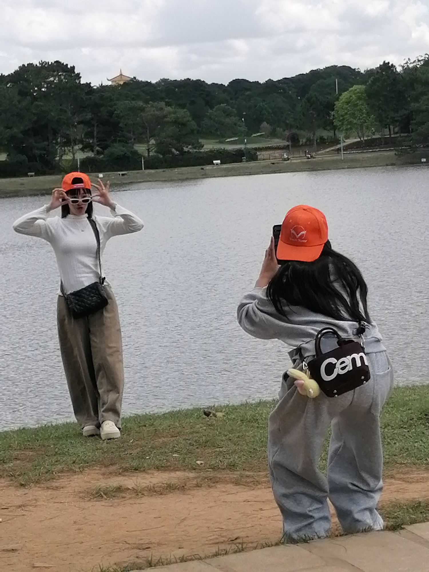 Two young women are posing for photos by a lake in Da Lat, Vietnam. The woman on the left, wearing an orange baseball cap and white sunglasses, is standing with her hands near her face, striking a pose. She is dressed in a white long-sleeved shirt and loose-fitting beige pants, with a black quilted crossbody bag. The woman on the right, also wearing an orange baseball cap and holding a smartphone, is in the process of taking a photo. She is wearing a grey hoodie and baggy grey sweatpants, and a small dark brown bag with "Cem" written in white letters is attached to her waist. Both women are standing on a grassy embankment overlooking the water. In the background, a dense forest lines the far side of the lake, with a distant building visible on a hilltop, suggesting the location is a park or a scenic spot within Da Lat. The sky is overcast.