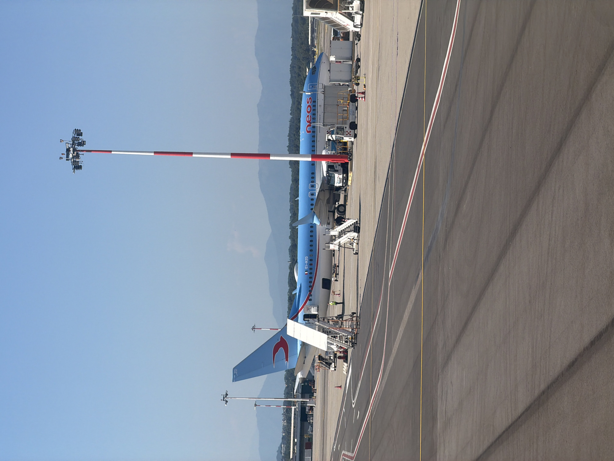 An airplane with "neos" written on its side is parked on the tarmac at Malpensa Airport in Ferno, Italy. The plane, which is blue and white, has a red logo on its tail. A set of stairs is connected to the plane, and a person in a yellow vest is standing near the plane. Several vehicles, including a tug and a baggage cart, are also on the tarmac. In the background, there are mountains and trees. The sky is clear and blue.