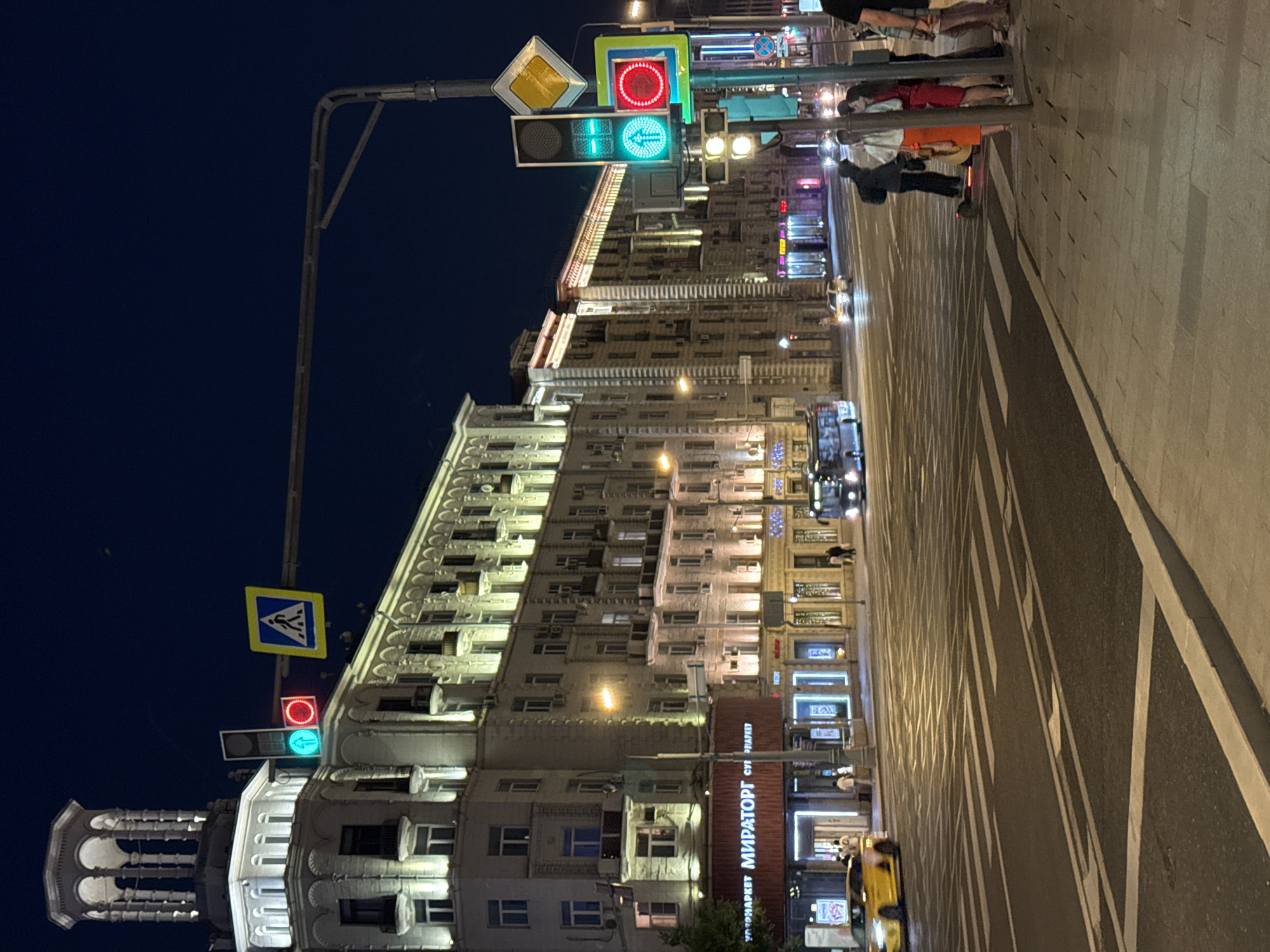 This image depicts a night scene on Novy Arbat street, 17, Moscow, Russia. The sky is dark, illuminated by various artificial light sources.
On the left, a multi-story, light-colored building in a classical architectural style features a distinctive decorative turret at its corner. The building's facade and windows are illuminated. The ground floor of this building houses a "МИРАТОРГ" (MIRATORG) supermarket, identifiable by its red illuminated sign, with several individuals visible near its entrance.
The foreground shows a wide asphalt road with multiple lanes, marked by white pedestrian crossing stripes and other road markings. A yellow car, likely a taxi, is visible parked or moving slowly near the left curb. Further down the street, on the right side of the road, a blue bus with illuminated headlights is present. Other vehicles with activated headlights are visible in the distance.
On the right side of the image, a group of approximately five to six pedestrians stands on a paved sidewalk, positioned near a utility pole. This pole supports multiple traffic signals and road signs. One vehicle traffic light displays a green signal, while another displays red. A pedestrian traffic light also shows a green signal, indicating "walk." Above these, a blue square sign with a white walking person (pedestrian crossing) and a yellow diamond-shaped sign (priority road) are attached to the pole. One person in the pedestrian group appears to be on a motorized scooter or similar device, positioned near the curb. The group includes individuals dressed in various casual attire, with some appearing to interact.