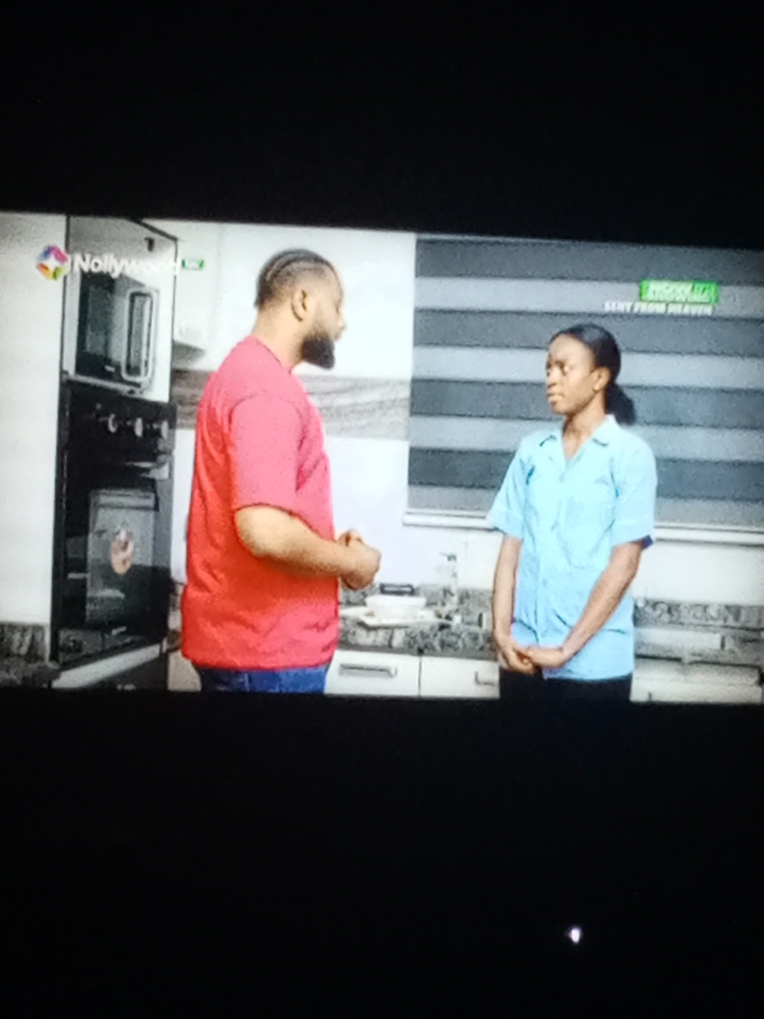 A scene from a Nollywood film entitled "Sent From Heaven" shows a man and a woman in a kitchen. The man is facing left, wearing a red shirt and blue jeans. The woman is facing right, wearing a light blue shirt and dark pants. A microwave and oven unit is to the left, and a window with horizontal blinds is behind the woman. The location is unknown.