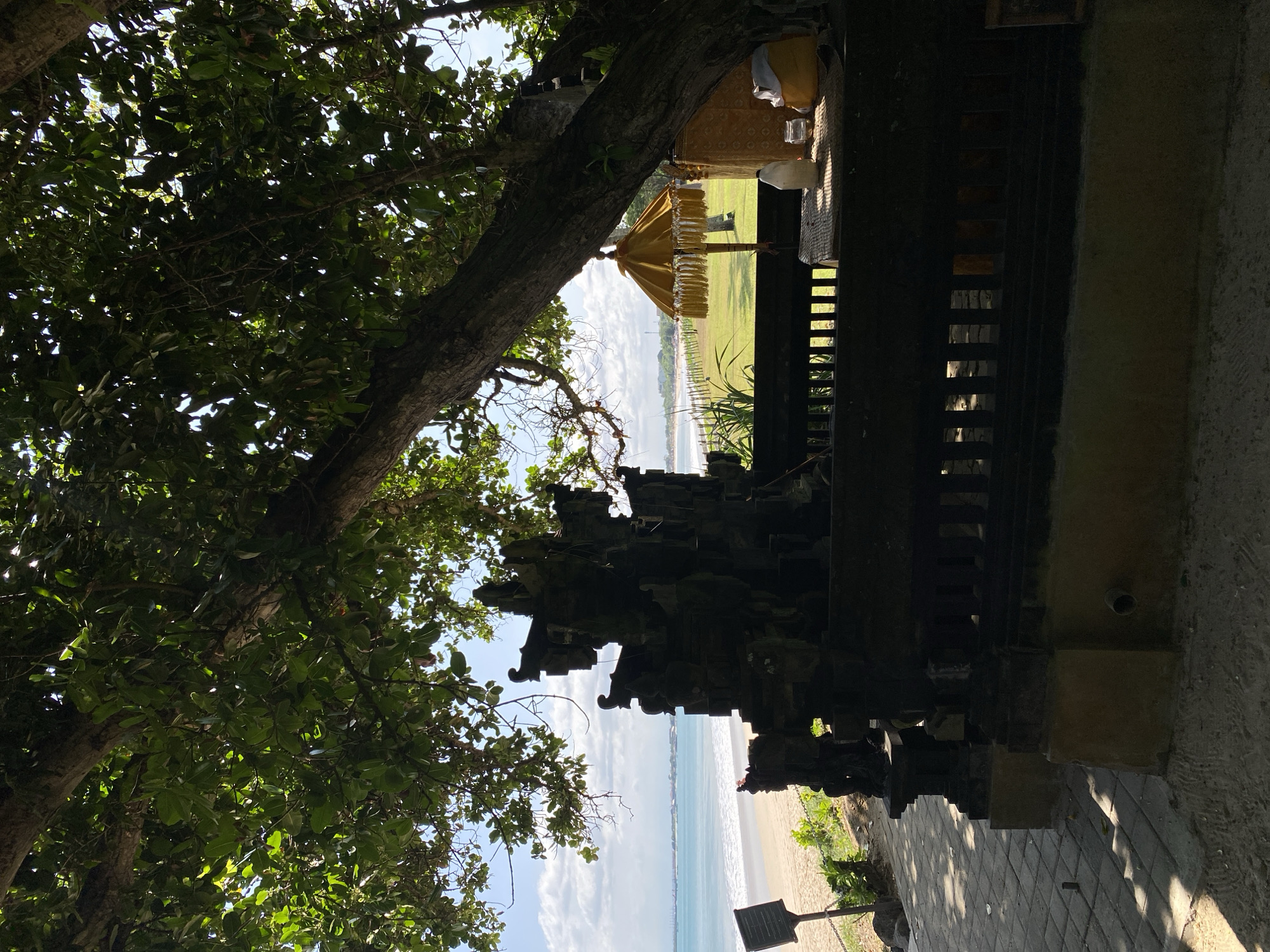 The image depicts a coastal scene in Kuta Selatan, Indonesia. The view is partially obscured by a tree with dense foliage in the foreground. Adjacent to the tree is an intricately carved, dark-colored structure, likely part of a temple or traditional building. Through the gaps in the structure, a view of the beach and ocean is visible. In the distance, a yellow ceremonial umbrella stands on a green lawn. The sky is blue with scattered clouds. The ground near the structure is paved.