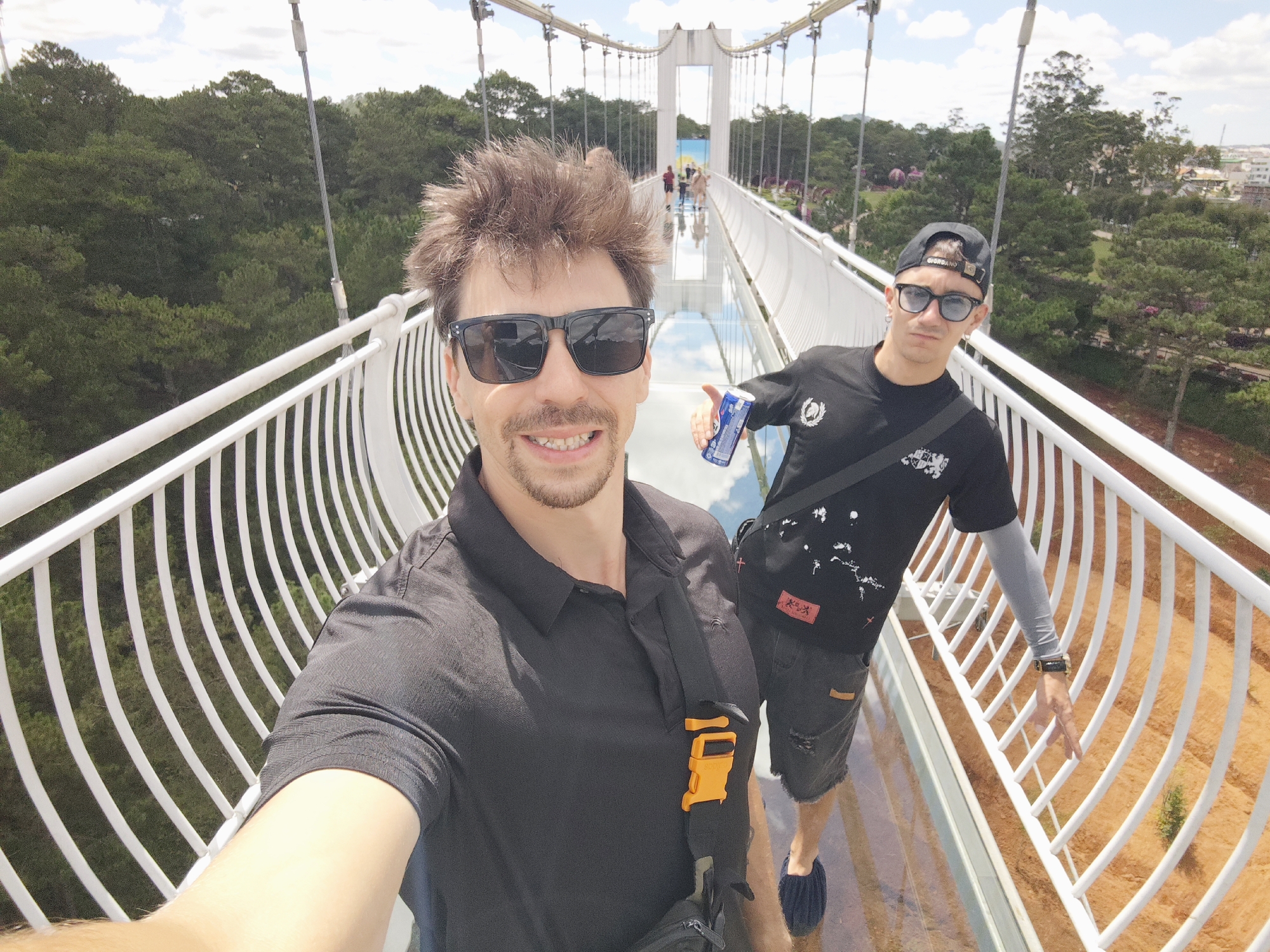 Two young men are taking a selfie on a transparent bridge in Da Lat, Vietnam, specifically on the Phuong Hoang bridge, also known as the Glass Bridge, located in Lam Vien Highland. The man in the foreground is wearing a black polo shirt, sunglasses, and a black crossbody bag with an orange buckle. He is smiling and holding the camera out for the selfie. The man in the background is wearing a black t-shirt with a graphic print, a black baseball cap, sunglasses, and a light gray arm sleeve. He is holding a blue can. The bridge has white railings and a glass walkway that offers a view of the orange earth and green trees below. In the background, other people can be seen walking on the bridge and enjoying the scenery. The sky is partly cloudy, with scattered white clouds against a blue sky. The surrounding area is covered with lush green trees, characteristic of the mountainous landscape of Da Lat. The overall impression is one of a tourist attraction offering a unique perspective of the natural beauty of the region.