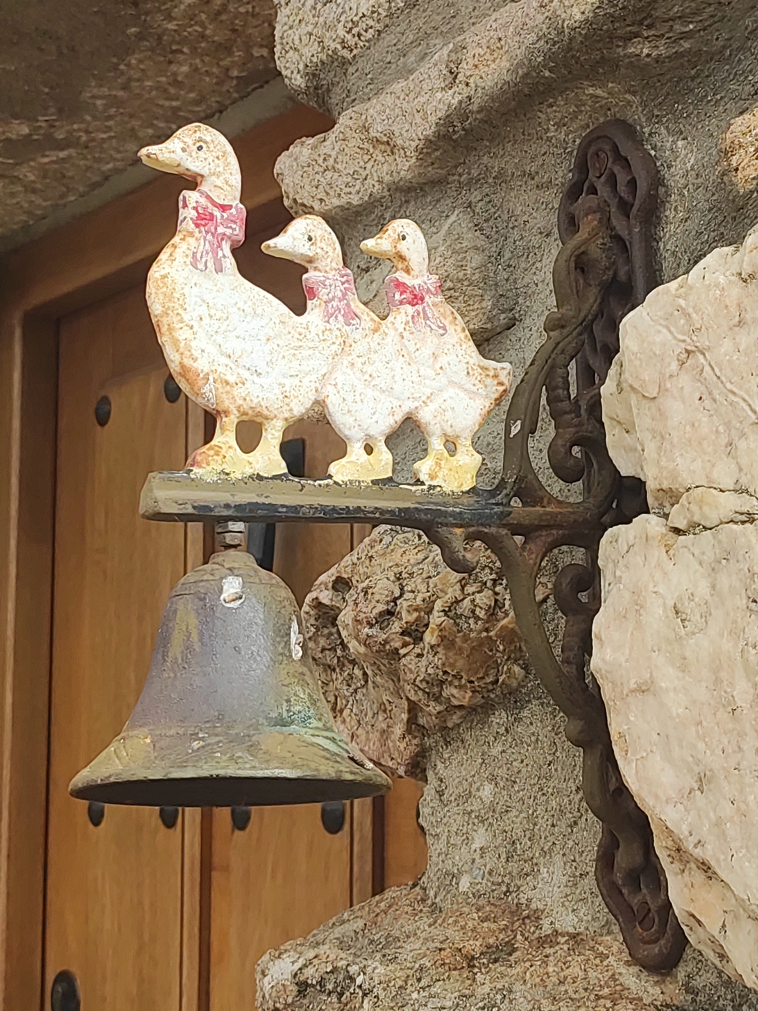 The image shows a decorative bell mounted on a stone wall next to a wooden door in the town of Sobradillo, Spain. The bell is made of metal and has a distressed, aged appearance. Above the bell is a decorative wrought iron bracket that supports three ceramic duck figures. The ducks are painted white with yellow feet and wear pink ribbons around their necks. The background consists of rough stone textures, suggesting an exterior architectural feature.