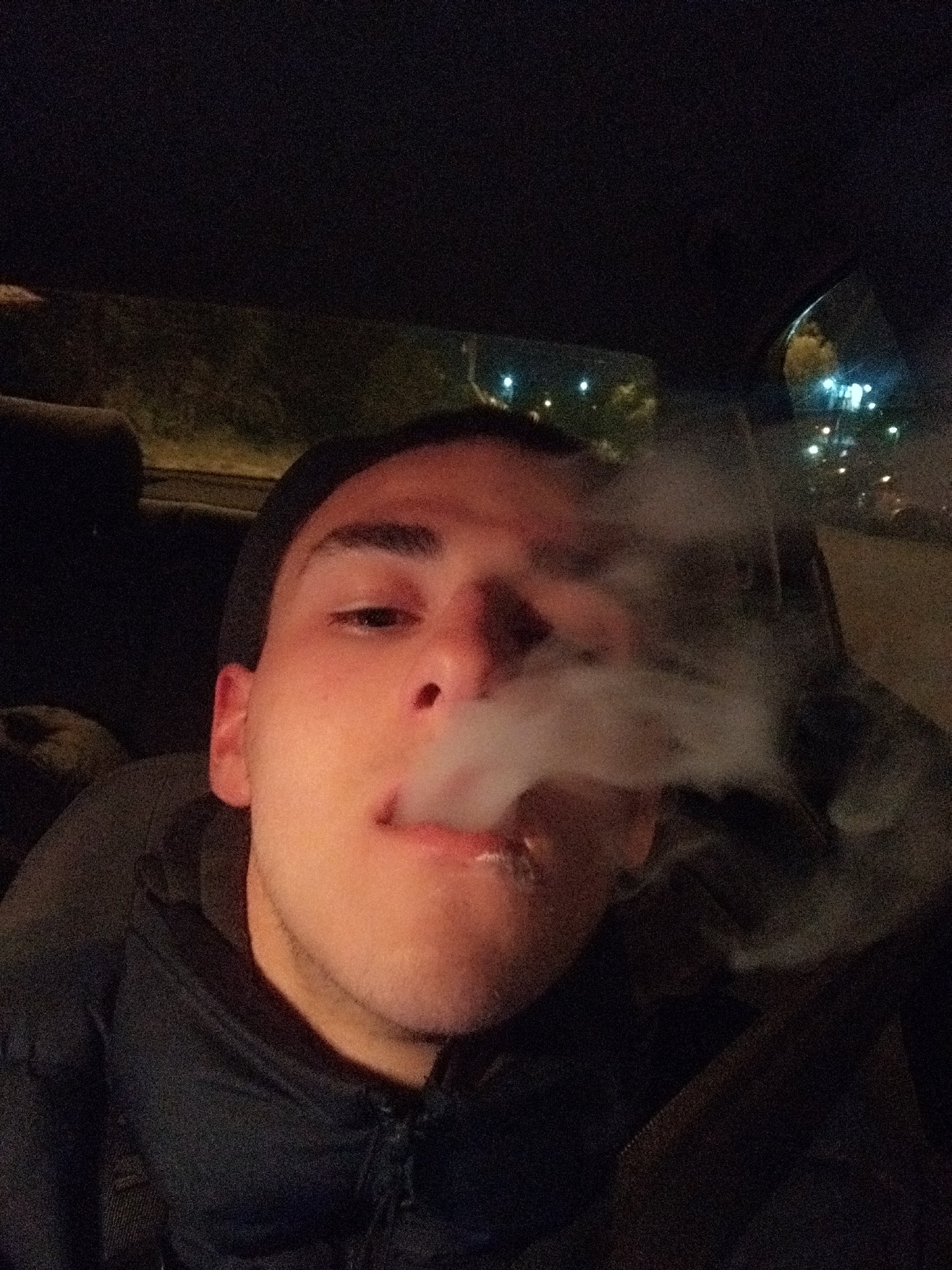 A low-angle, close-up shot depicts a young male inside a vehicle at night. The man is exhaling a cloud of smoke. He has dark hair, light skin, and is wearing a dark jacket or hoodie. The background is dark, with blurred lights visible through the car windows. The lights suggest an urban environment. I cannot determine the location in Sofia with this information.