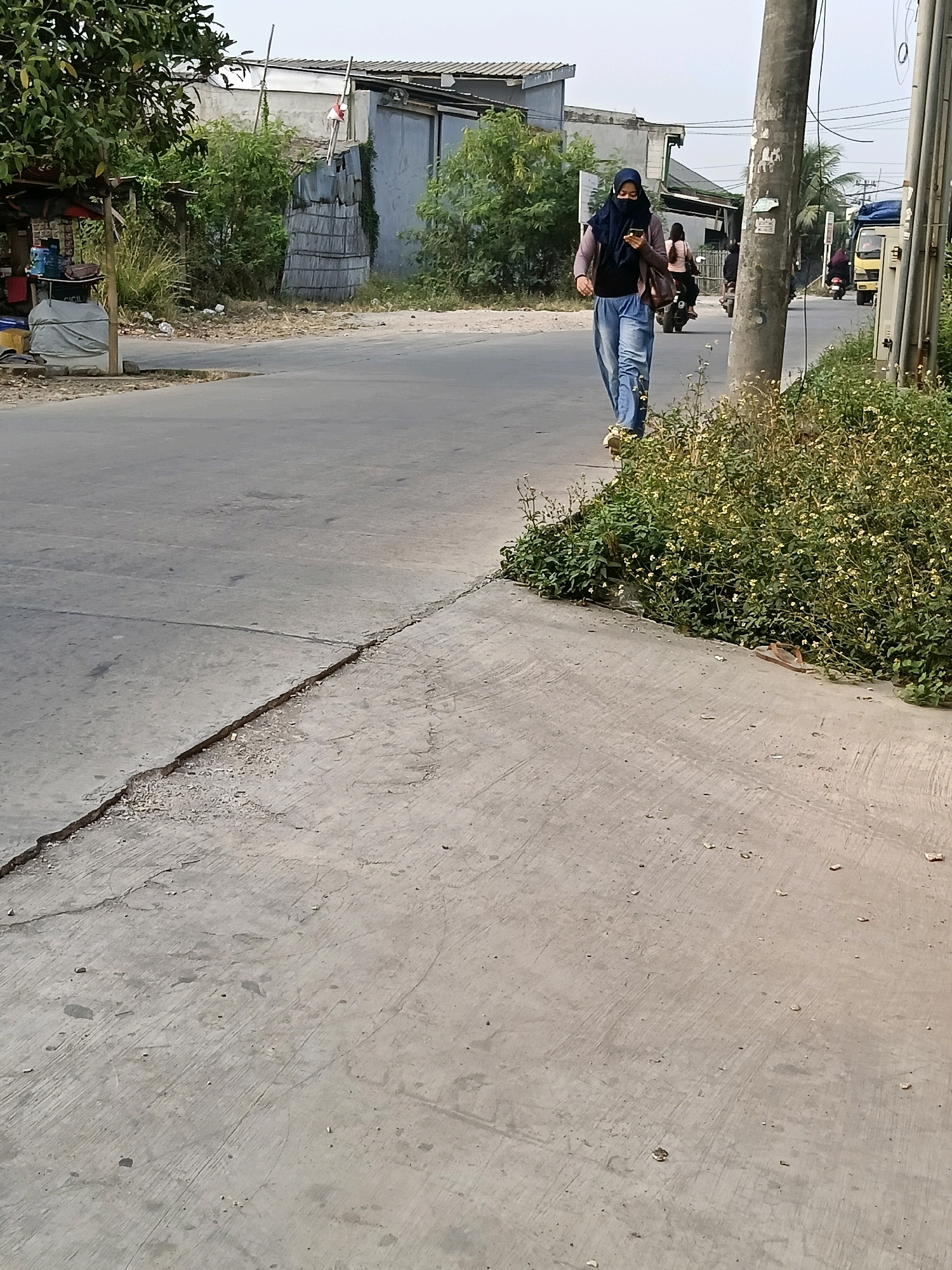 A woman wearing a dark blue hijab and a light pink long-sleeved top over a black shirt, along with blue jeans, walks on the side of a paved road in the city of Belimbing, Indonesia. She is looking at her phone, which she holds in her left hand. Her right hand is at her side. She is walking towards the viewer. To her right, a tall, rough-textured utility pole is visible, with wires extending from it. Beyond the woman and the pole, several other people are on motorcycles, and a yellow truck is driving away from the viewer. The road is flanked by a sidewalk made of concrete that is cracked in several places. Dense green foliage with small white flowers grows along the edge of the sidewalk. On the left side of the road, there is a small roadside stall with a canopy, displaying several bottles of water and other items. Behind the stall, there are green trees and buildings. The sky is overcast.