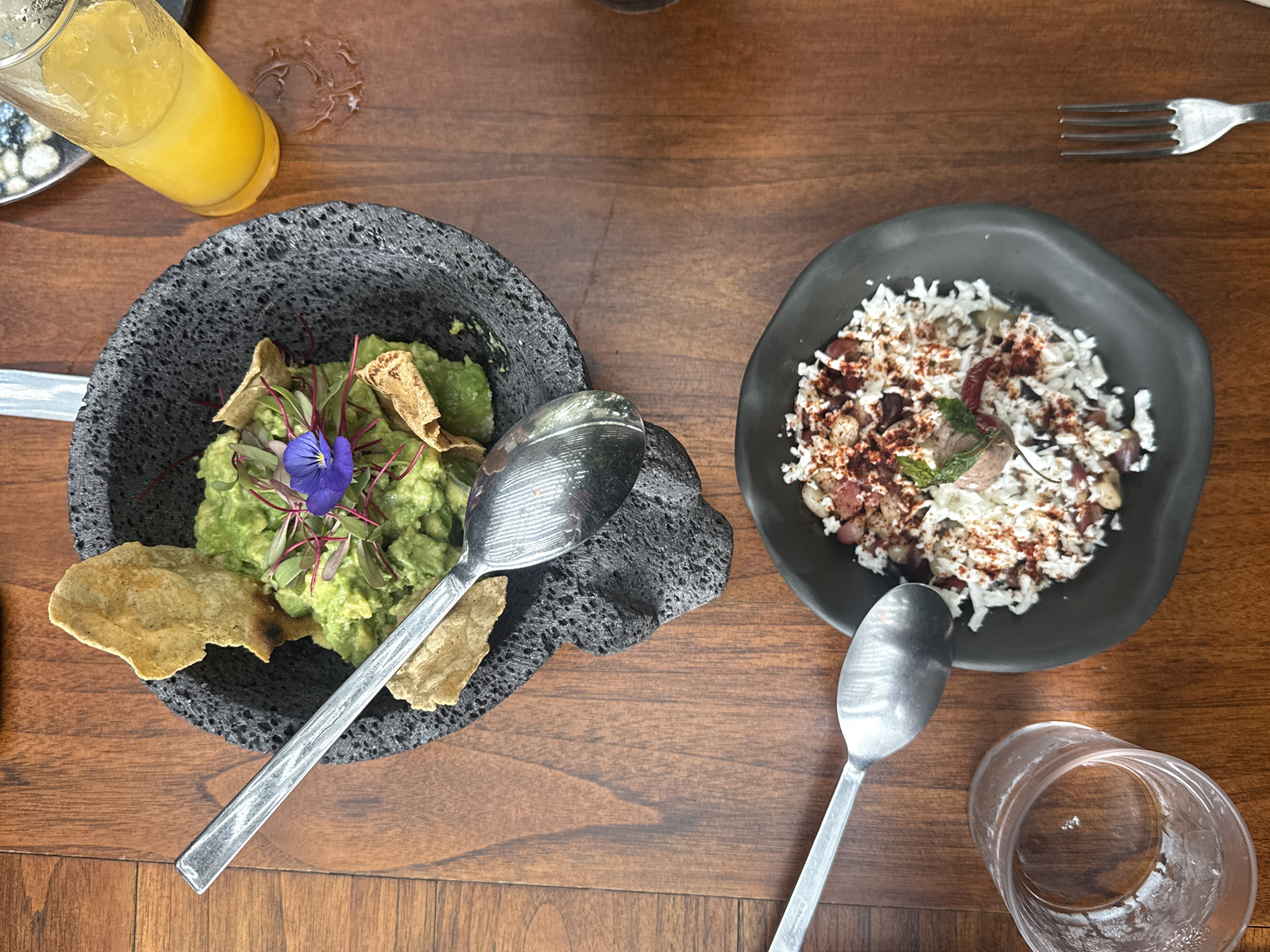 The scene depicts two dishes and beverages on a wooden table, likely in a restaurant in Mexico City, Mexico. On the left is a black molcajete filled with guacamole, topped with edible flowers and tortilla chips. A silver spoon rests on the side. Next to it is a glass of orange juice. On the right, a black plate contains a salad-like dish with white cheese, beans, and chili powder. A silver spoon is placed next to the plate, and an empty glass is near the bottom edge of the frame. A fork is visible in the upper right corner. The table has a visible wood grain pattern. The lighting suggests an indoor environment.