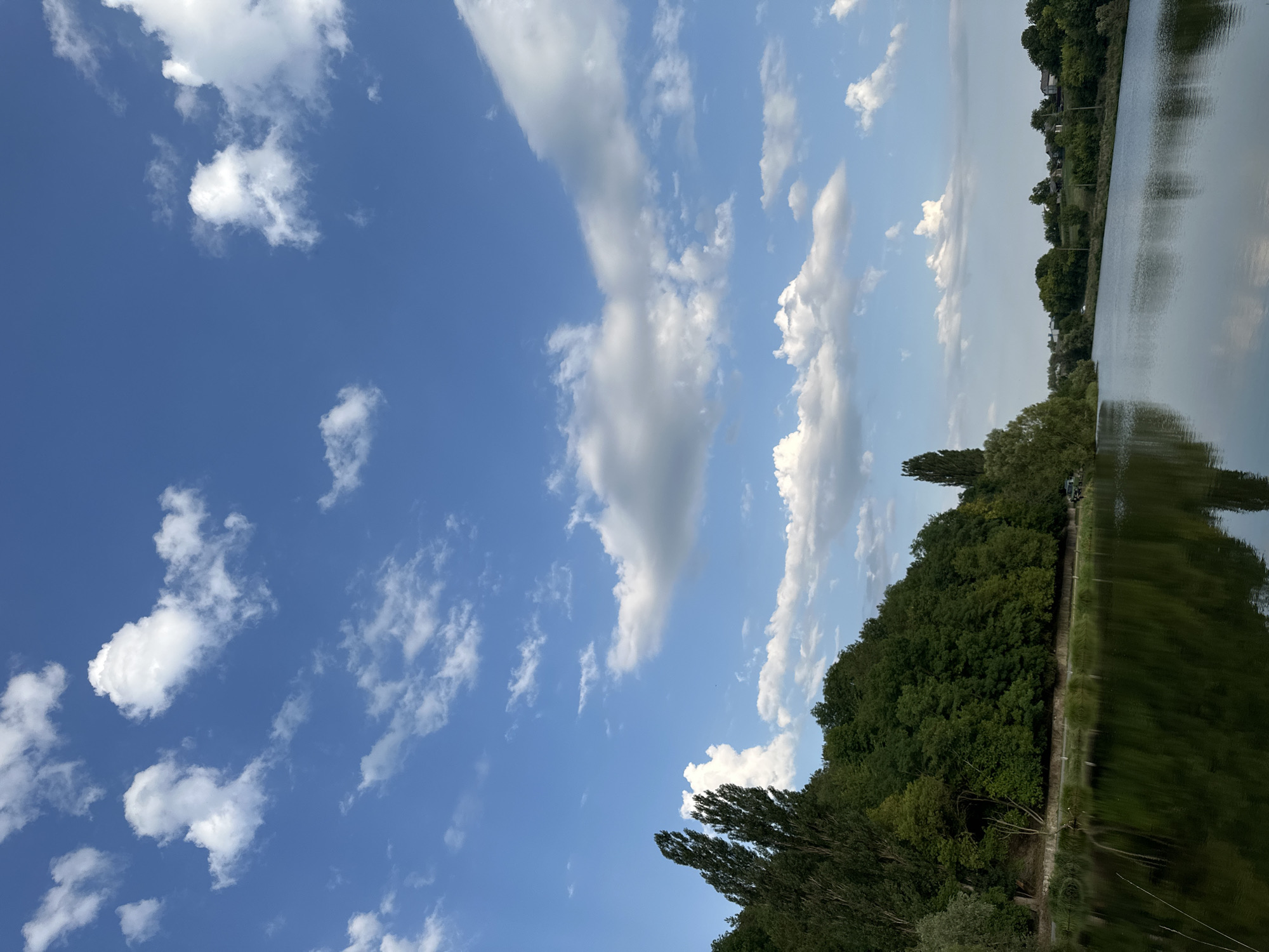 The image depicts a serene natural landscape with a body of water in the foreground, reflecting the sky and surrounding trees. A dense forest lines the left bank of the water, with a winding path visible along its edge. The sky above is a vibrant blue, dotted with fluffy white clouds. In the distance, the right bank of the water is lined with trees and houses, suggesting a rural or suburban area. The overall impression is one of tranquility and natural beauty. This scene is consistent with the natural environment found in and around the village of Makiv, Ukraine, which is known for its picturesque landscapes and bodies of water.