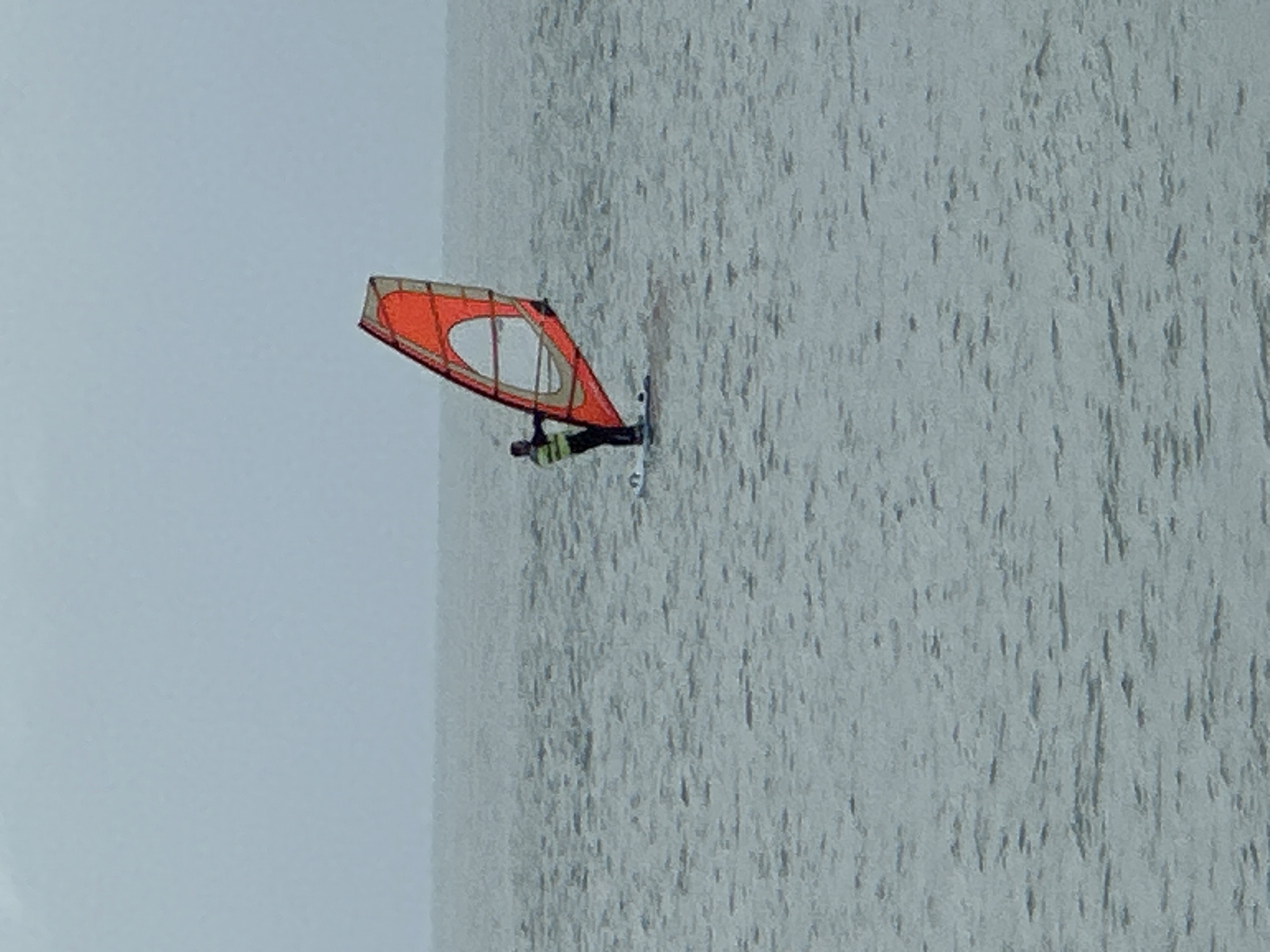 A windsurfer is seen on the water in Sochi, Russia. The water is grey and choppy, and the sky is overcast. The windsurfer has an orange sail with a white circle in the middle. The person on the windsurfer is wearing a wetsuit and a helmet.