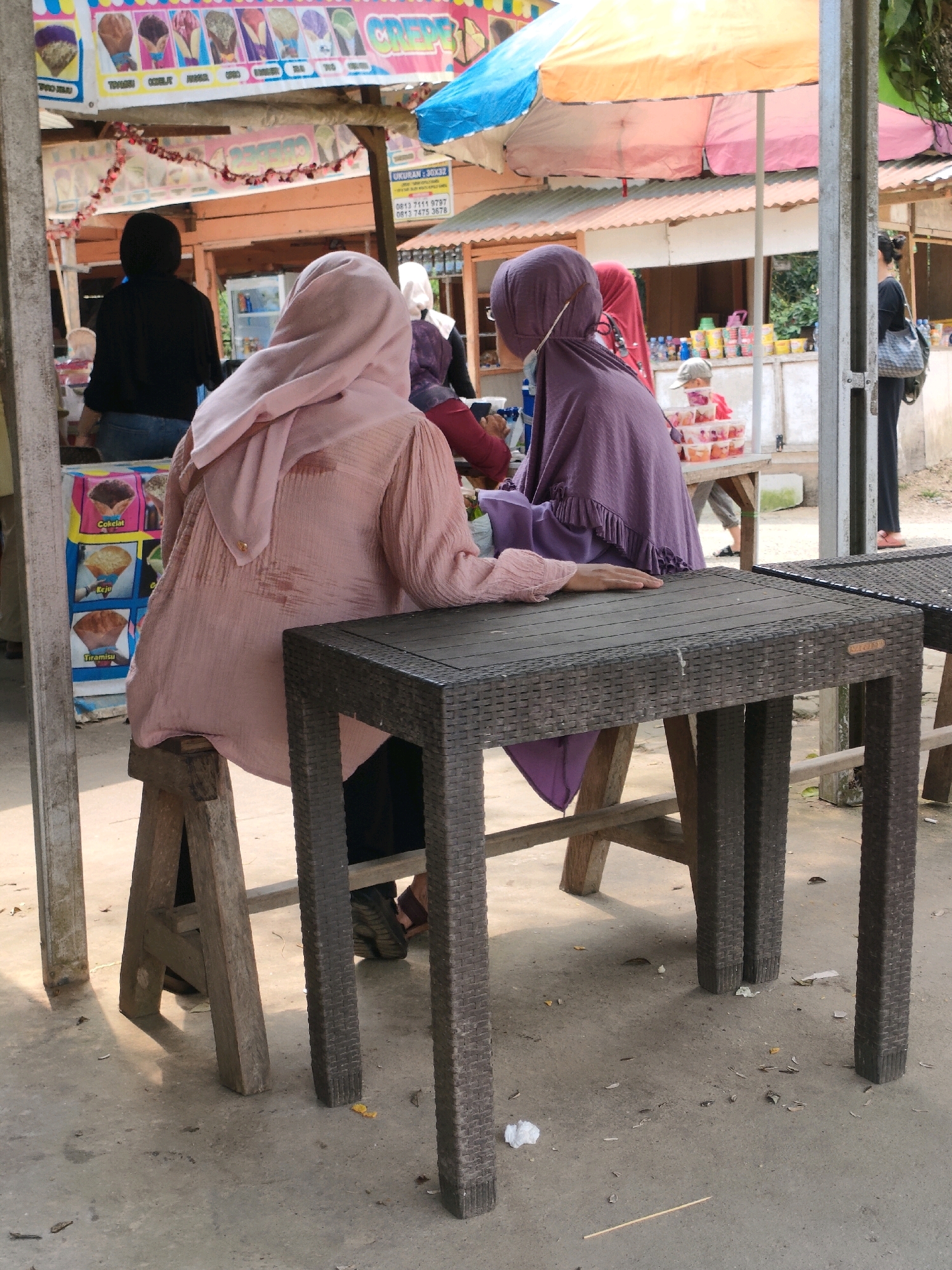 Two women wearing hijabs are seated at a small, dark brown, rattan-style table. One woman, on the left, is wearing a light pink hijab and a light pink, long-sleeved, textured shirt. She is seated on a wooden stool and is looking down at the table, with her right hand resting on it. The second woman, on the right, is wearing a dark purple hijab and a dark purple, ruffled garment. She is seated on a chair and is facing away from the camera, partially obscured by the first woman and the table.
Behind them, a food stall is visible, with a sign displaying images of ice cream cones labeled "Coklat," "Keju," and "Tiramisu." Further back, other people are seen, including a person in a black top and jeans standing to the left, and another person in black, wearing a backpack, standing to the right. The environment appears to be outdoors, with a concrete ground, sunlight casting shadows, and a corrugated metal roof visible in the background. The scene is likely a street food vendor or a small market stall.