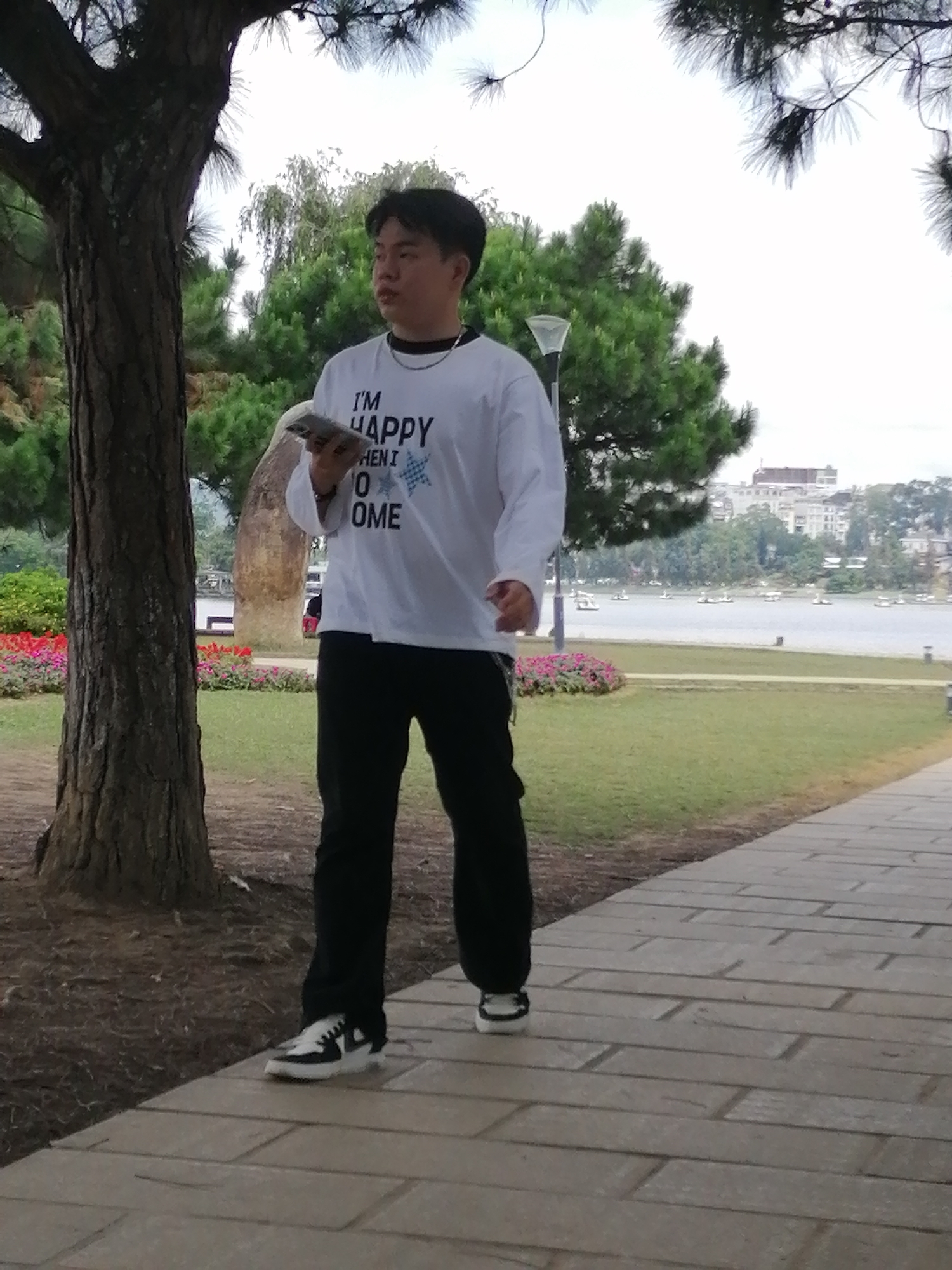 A young man walks along a paved path beside Xuan Huong Lake in Da Lat, Vietnam. He is wearing a white long-sleeved shirt with a graphic print that reads "I'M HAPPY WHEN I COME HOME", black pants, and black and white sneakers. He holds a phone or small book in his left hand. The background features the lake with paddle boats, lush green trees, and buildings in the distance, suggesting the urban fringe of Da Lat. The path is made of light-colored rectangular stones, and to the left of the path, there is a tree with dark bark and pine needles, and a flower bed with pink and red flowers. The weather appears overcast.