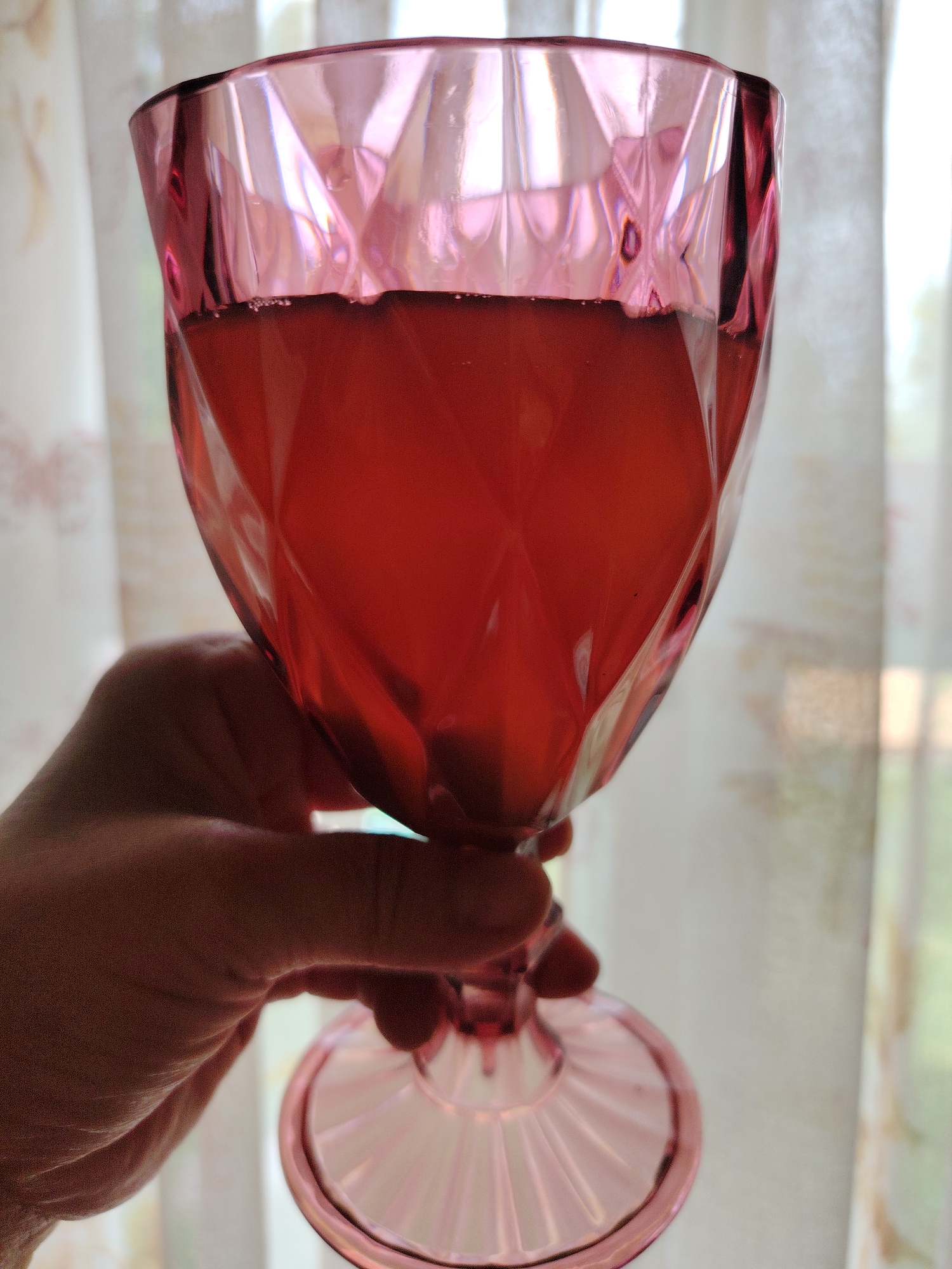 A hand holds a pink, faceted glass goblet filled with a red liquid. The goblet is angled upwards, and the liquid fills about two-thirds of its volume. The background consists of sheer curtains, illuminated by natural light, suggesting an indoor setting. The scene is devoid of any specific landmarks or architectural elements that would indicate a location in the city of Кривец, Russia.
