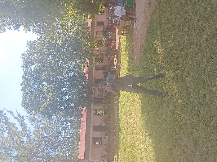 An outdoor scene in Idakho South ward, Kenya, features a male subject in a dark suit playing a dark wind instrument in the foreground of a grassy area. His hands are positioned to operate the instrument at mouth level. In the background, a single-story building with a reddish-brown corrugated iron roof and light-colored walls is visible, fronted by a seated and standing audience, largely composed of children. Trees with green foliage line the background, and the sky is clear. Direct sunlight casts shadows across the scene.