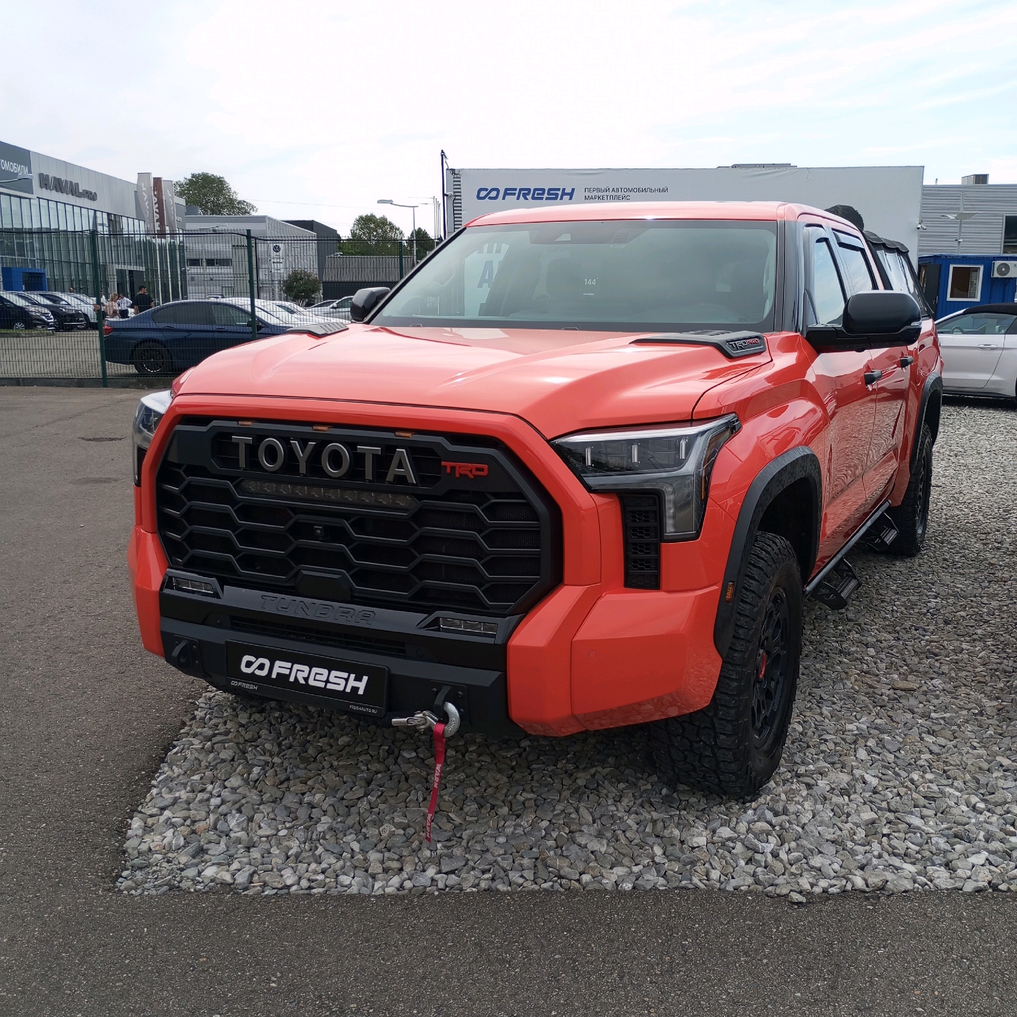 An orange Toyota Tundra TRD Pro truck is parked on a gravel lot in front of a dealership in Sochi, Russia. The truck's front grille has "TOYOTA" emblazoned across it in silver lettering, with a red "TRD" logo to the right. A black bumper with "TUNDRA" written on it is visible below the grille. A red tow strap is attached to a hook on the bumper. The dealership building in the background has signs for "HAVAL" and "CO FRESH". Several other vehicles, including a dark blue sedan and a white convertible, are parked in the lot behind the Tundra. The sky is overcast.
