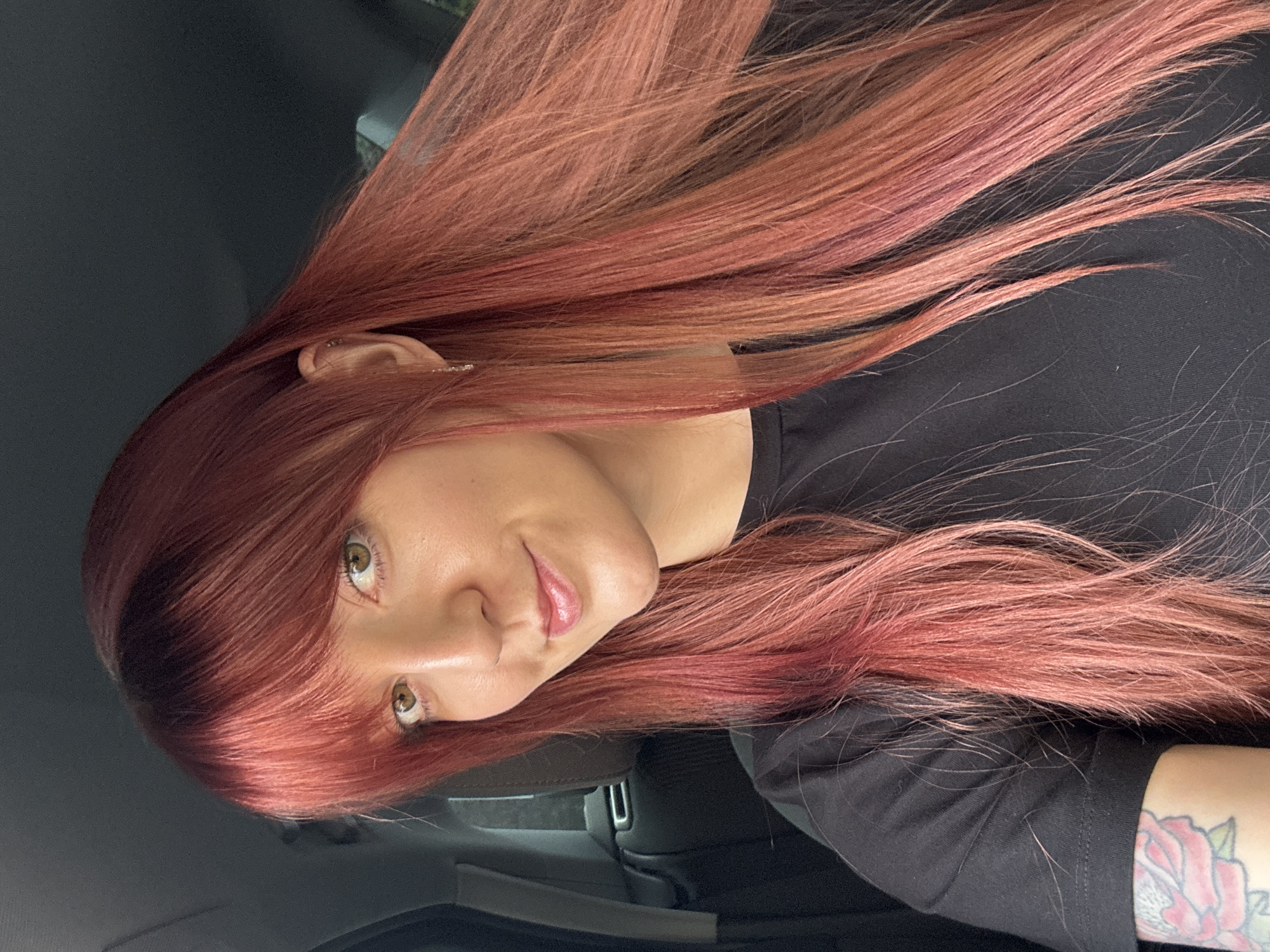 A woman with auburn, shoulder-length hair and fair skin is facing the camera, looking to her left. She is wearing a black t-shirt. A portion of a tattoo, a rose with green leaves, is visible on her left forearm. The background is dark, appearing to be the interior of a car. There is no information in the image to suggest the location is in the city of Izhevsk, Russia.
