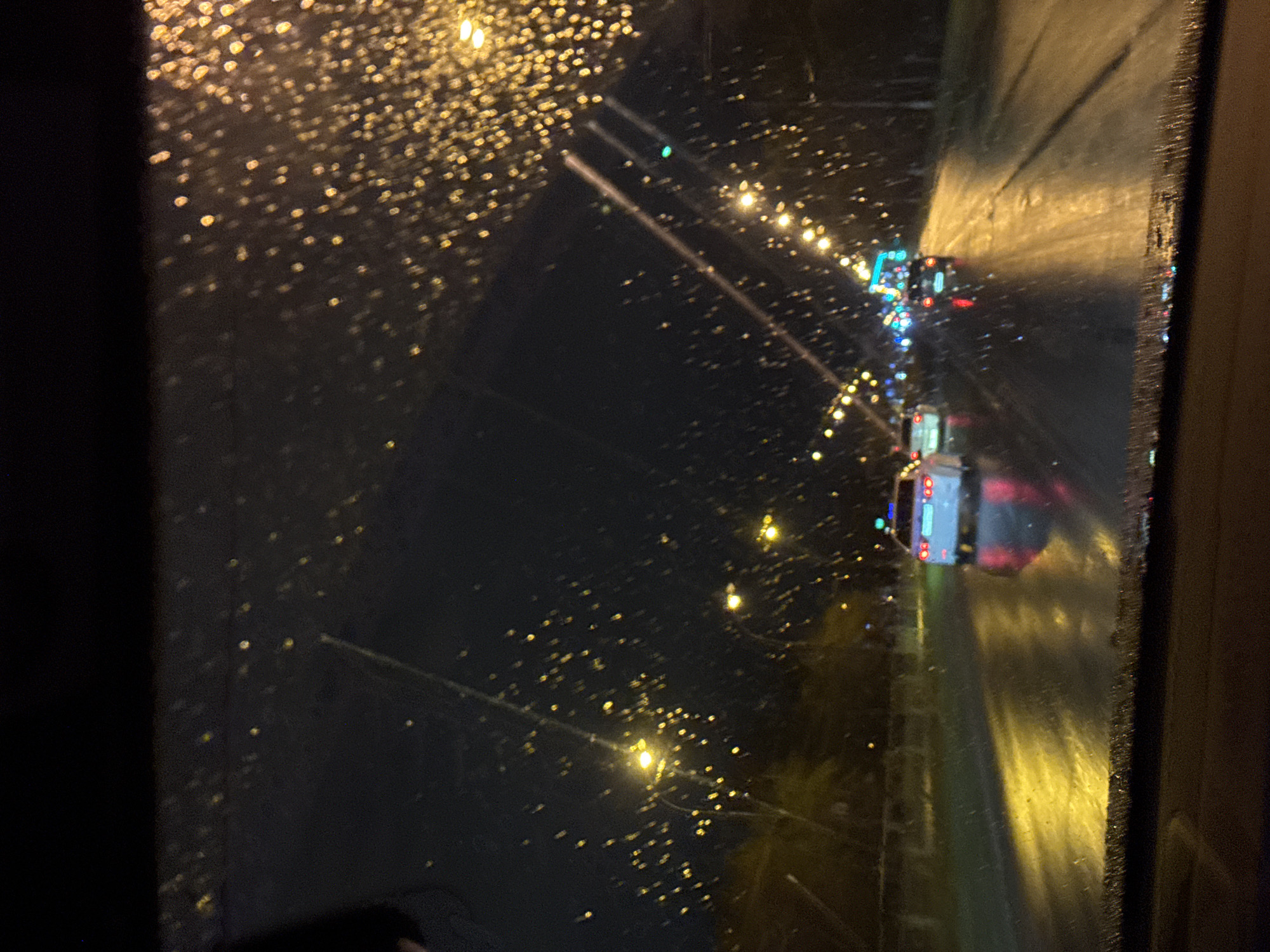 The image captures a nighttime scene viewed through a window, likely from a moving vehicle, with numerous water droplets distorting the view. The time of day is clearly night, characterized by the dark sky and prominent artificial lighting. The presence of water droplets on the window indicates rain or condensation, and the road surface appears wet and reflective, further suggesting recent or ongoing precipitation.
The primary subjects visible beyond the window are a road illuminated by streetlights and the lights of other vehicles. In the mid-ground, a white truck or bus is discernible, with its red taillights visible. Further down the road, more vehicle lights, including blue/white headlights and red taillights, indicate traffic. The streetlights line the road, casting warm yellow-orange glows that create significant bokeh effects due to the water on the window and the depth of field. The overall scene is blurred and atmospheric, with the water droplets transforming distant lights into soft, shimmering orbs.
The left side of the frame is dominated by a dark, out-of-focus area, possibly part of the vehicle's interior or a structural element. The image conveys a sense of observation from within a contained space, looking out at a bustling, wet nocturnal environment. There is no visible text in the image.