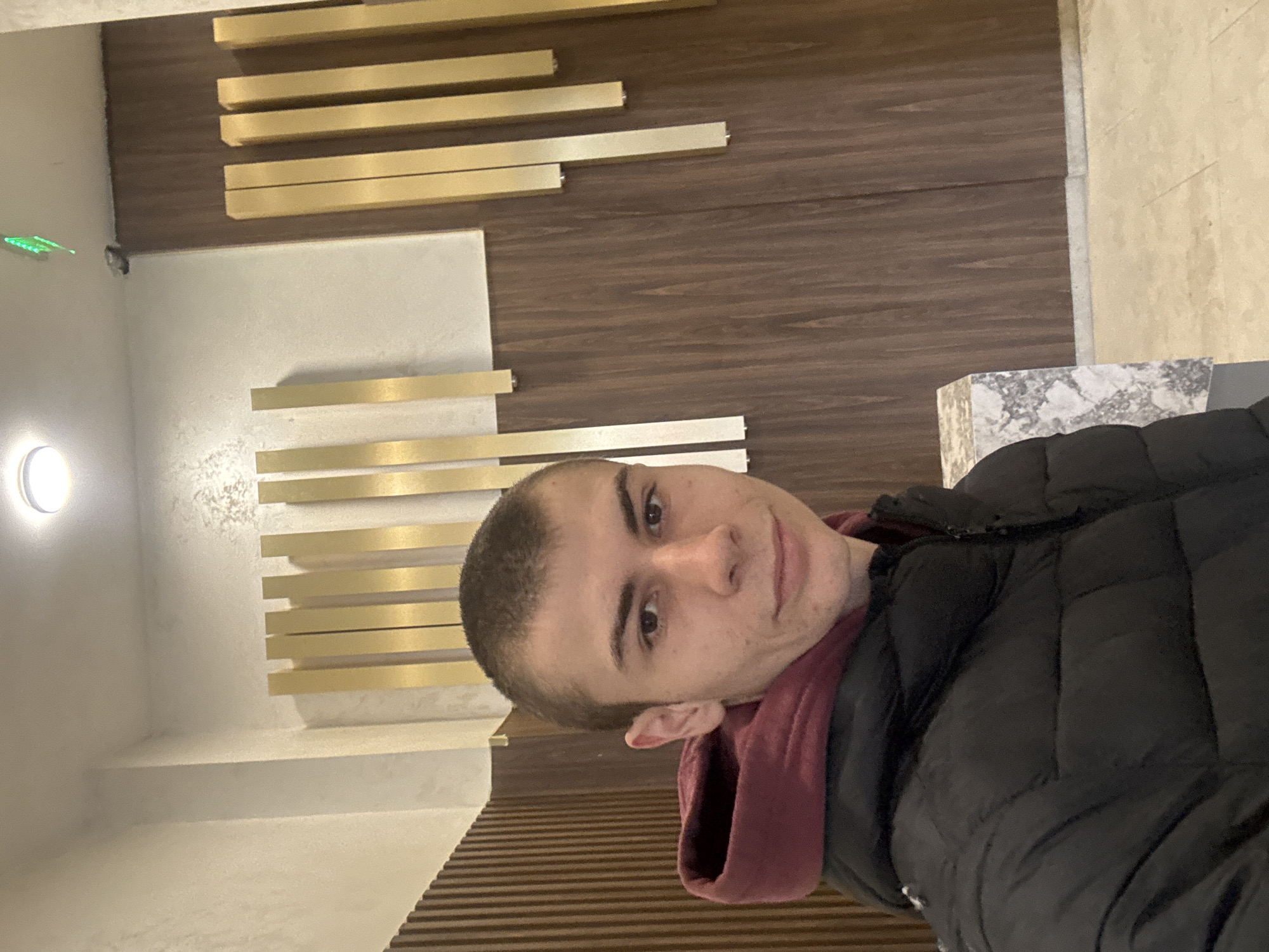 A medium shot depicts a person with short brown hair wearing a black puffy jacket and a maroon hoodie. The person is in a modern building. The background features a wall with horizontal gold bars against a light-colored surface and wood paneling. Based on the architectural style and the person's location, the image may have been taken in Plovdiv, Bulgaria, but this cannot be conclusively determined from the image alone.