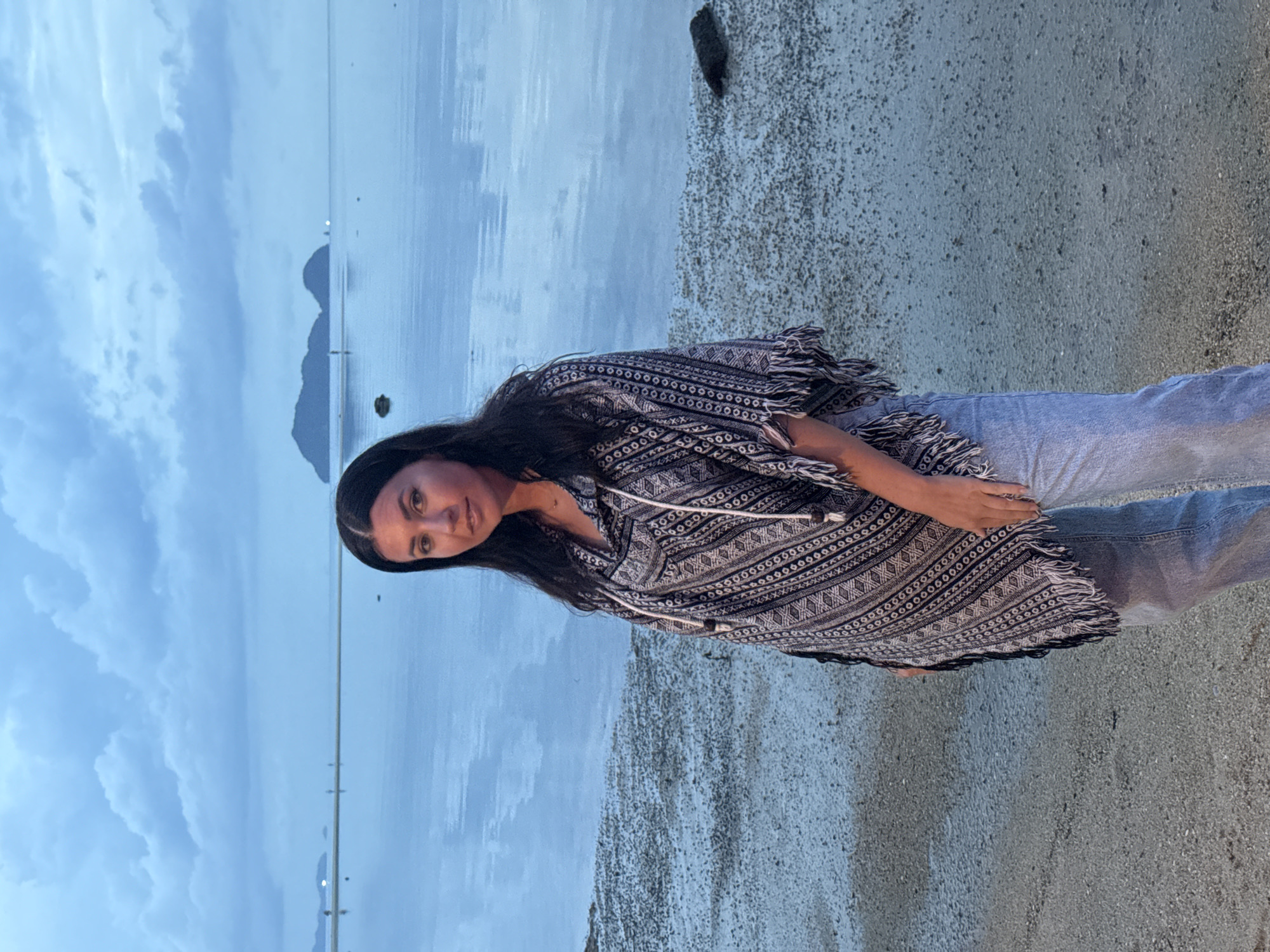 A woman with long, dark hair is standing on a sandy beach. She is wearing a gray and white patterned poncho and jeans. The beach is located in Baan Sri Thanu, Thailand. In the background, there is a body of water reflecting the sky. Mountains are faintly visible in the distance. The sky is overcast with blue-gray clouds.