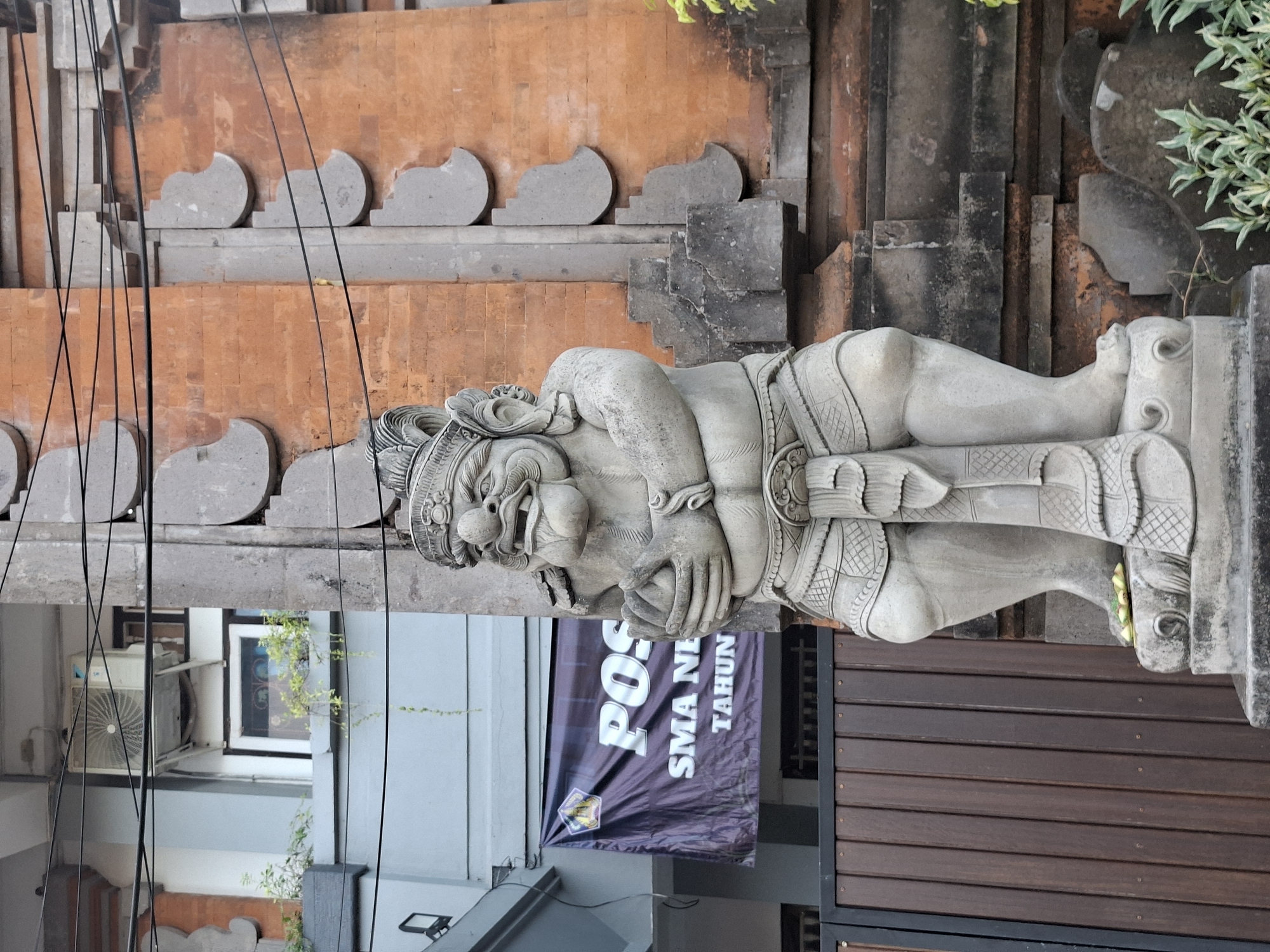 In Blahbatuh, Indonesia, a weathered stone statue of a Balinese guardian stands prominently in front of a brick building. The statue, with its intricately carved details, is positioned on a stone base. Behind the statue, a dark banner with white text reads "POS SMA NE TAHUN". The building's architecture features a terracotta brick facade adorned with gray stone accents. Overhead, a network of black electrical wires crisscrosses the frame. To the left of the statue, a section of a gray building is visible, along with a wooden fence and glimpses of greenery. An air conditioning unit is mounted on the exterior of the gray building.