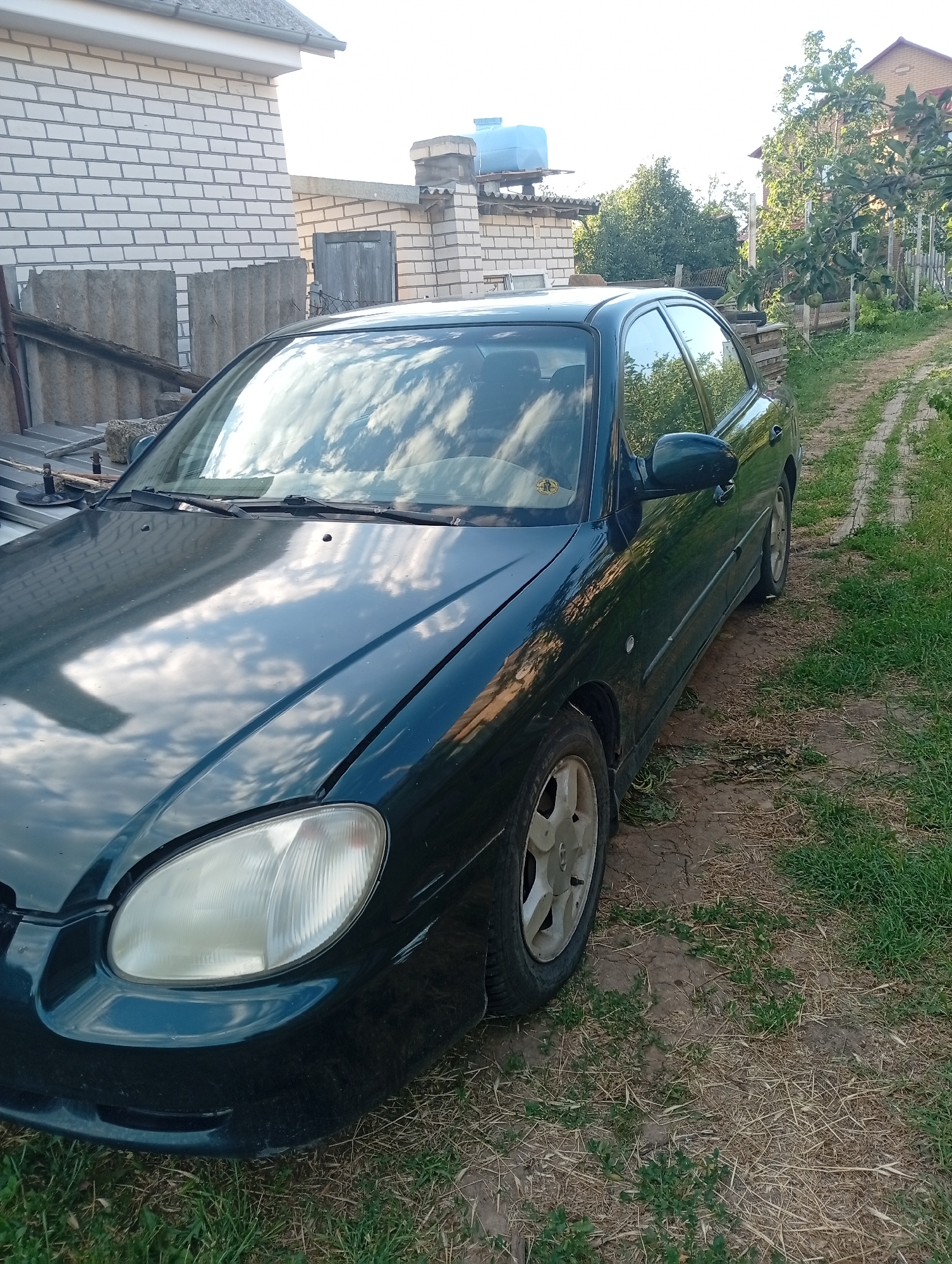 A dark green sedan is parked on a dirt path next to a grassy area. A white brick building with a gray roof is visible to the left of the car, and a fence with trees and foliage is to the right. The sky, reflected in the car's hood and windshield, shows white clouds. The car appears to be stationary in a rural or semi-rural setting.