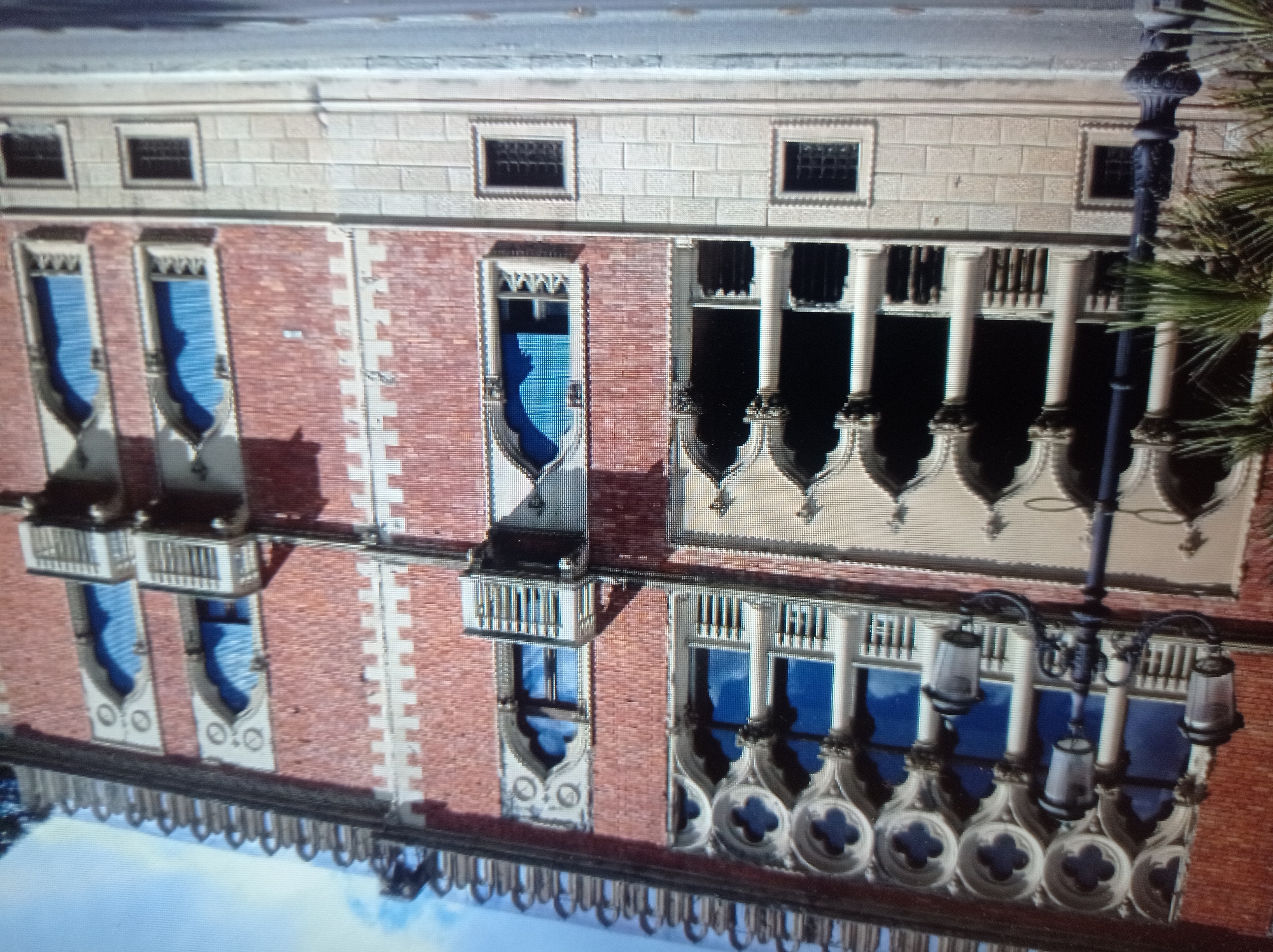 The image shows a close-up of a red brick building with ornate architectural details, likely from a historical period. The building features multiple arched windows with decorative tracery, balconies, and a distinctive crenellated parapet along the roofline. Some of the windows are covered by blue shutters. A black wrought-iron lamppost with multiple lights stands in the foreground on the left, partially obscured by palm fronds. The lower part of the building is faced with light-colored stone blocks, and there are small, dark-paned windows at ground level. The scene appears to be a street view, with a road in the foreground. There are no people or animals visible.