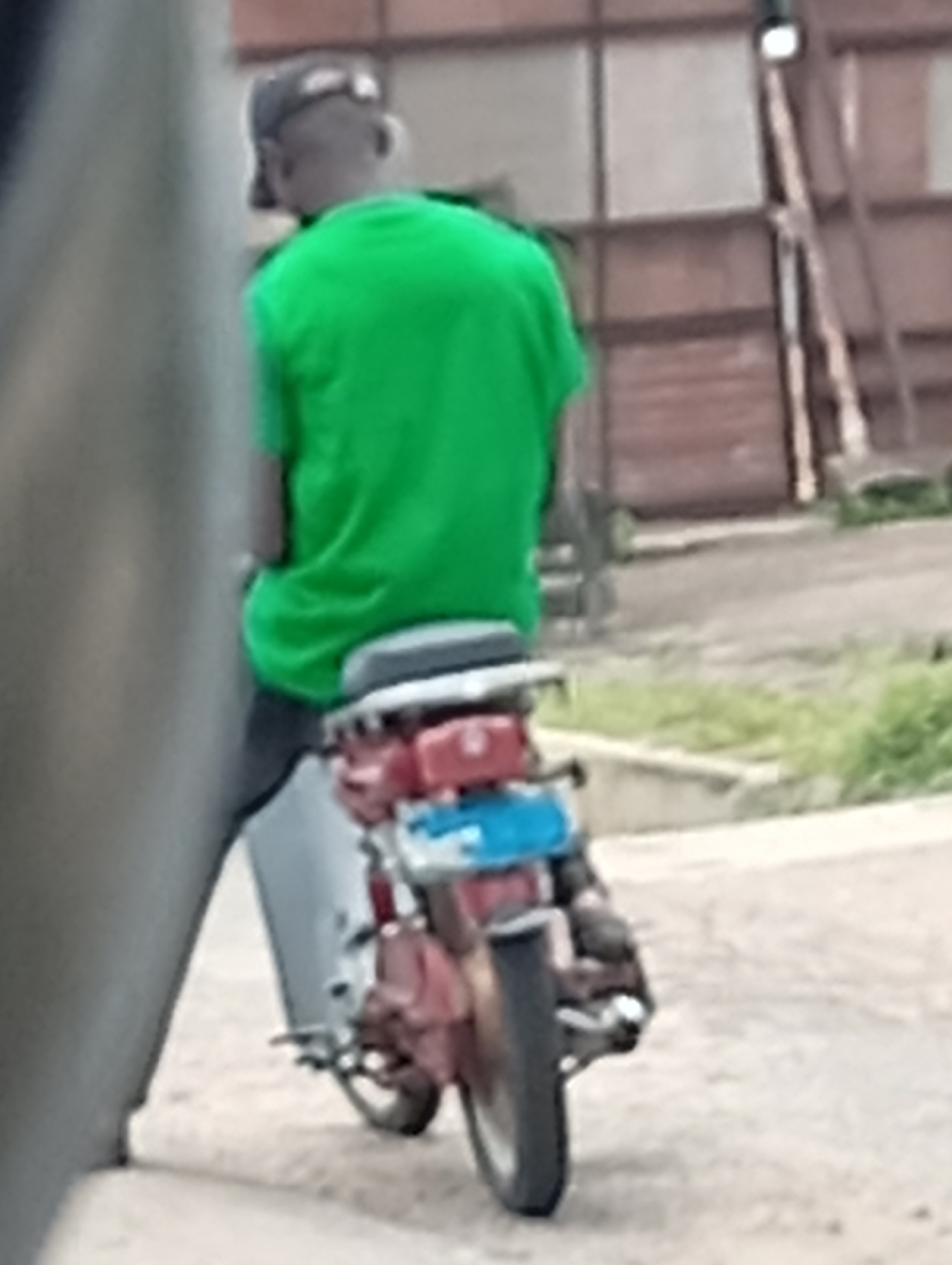 In an outdoor urban setting, a person wearing a green t-shirt and a baseball cap is seated on a red and blue motorcycle. The person's back is to the camera, and they appear to be looking forward. The motorcycle has a license plate on the back. The background consists of a corrugated metal wall and some overgrown grass. The scene is blurry and taken from a distance, suggesting it may be a candid shot.