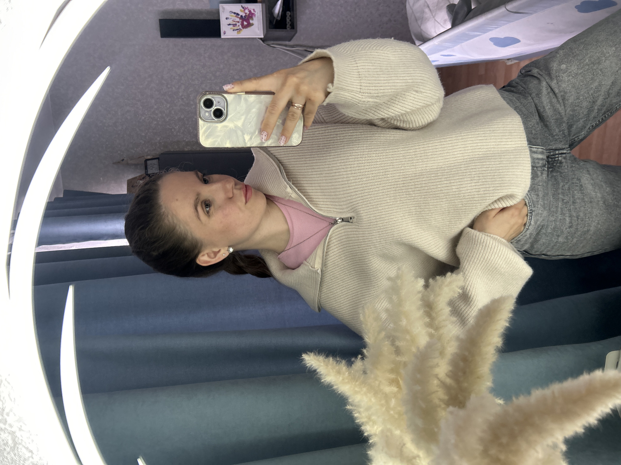 A young woman is taking a selfie in a mirror. She is wearing a pink t-shirt, a beige sweater with a zipper, and grey jeans. She is holding a white iPhone. The background includes a grey wall, a blue curtain, and a vase with pampas grass. Items on a shelf can be seen reflected in the mirror, including what may be a box set of K-Pop group BTS. The location appears to be indoors. There is no evidence the image was taken in the city of Klin, Russia.