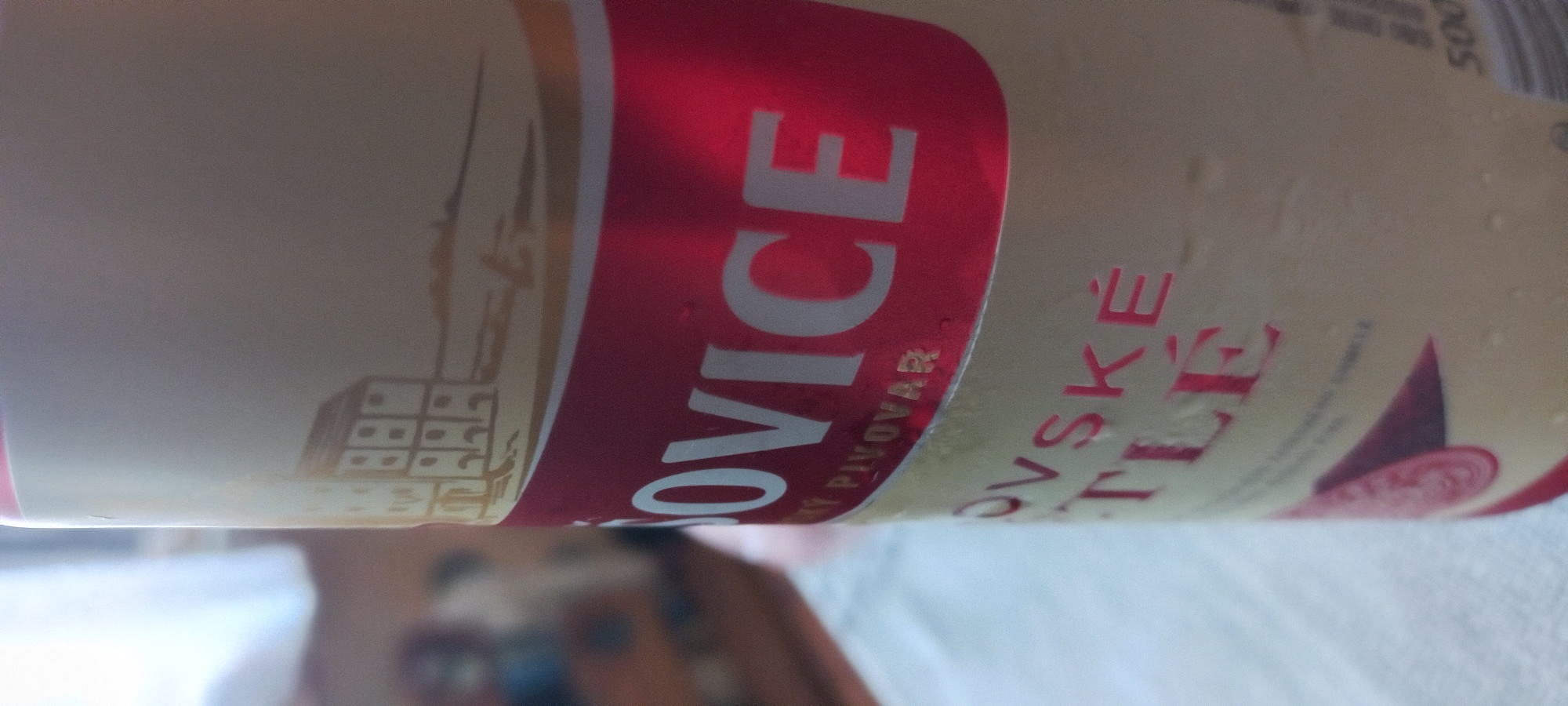 A close-up, angled view captures the upper portion of a cold beverage can in an indoor setting in Kryvyi Rih, Ukraine. The can, cylindrical and light cream-colored, features a prominent red horizontal band with white lettering that reads "OVICE". Above this band, in smaller gold letters, is "SKÝ PIVOVAR". An illustration of a multi-story, European-style building with visible windows, rendered in a monochrome golden-brown, is depicted above the red band on the can's cream background, positioned on a low horizon line with indistinct landscape features. Below the red band, more red text is partially visible, reading "OVSKÉ" and "TLÉ". Condensation in the form of small water droplets covers the surface of the can, indicating a temperature differential with the ambient environment. The very bottom right of the can shows "500", likely denoting a volume of 500ml. The background is significantly blurred, exhibiting a shallow depth of field. To the left, a bright, diffuse light source, possibly a window, creates a soft illumination. Indistinct, blurry shapes suggestive of furniture or other objects are visible in the far background. No individuals are clearly discernible.