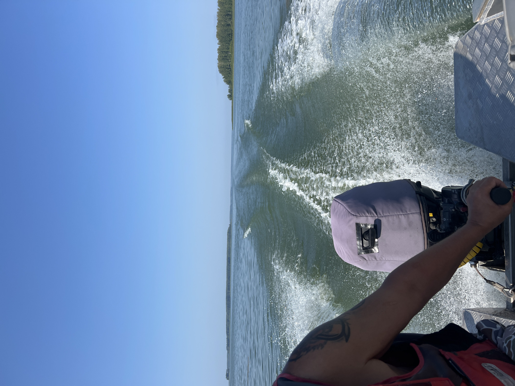 A first-person perspective shot from a moving boat on a sunny day. The viewer is looking out across a body of water, leaving a wake behind. In the foreground, to the left, is the arm and shoulder of a person wearing a red and black life vest. The person's arm has a tattoo on it. To the right of the frame, the back of a gray outboard motor cover is visible. The water is calm with ripples and white foam from the boat's movement. In the distance, a tree-covered island or peninsula stretches across the horizon under a clear blue sky. There are no buildings or other prominent man-made structures visible in the image. The location is identified as Beregovoe, Russia.