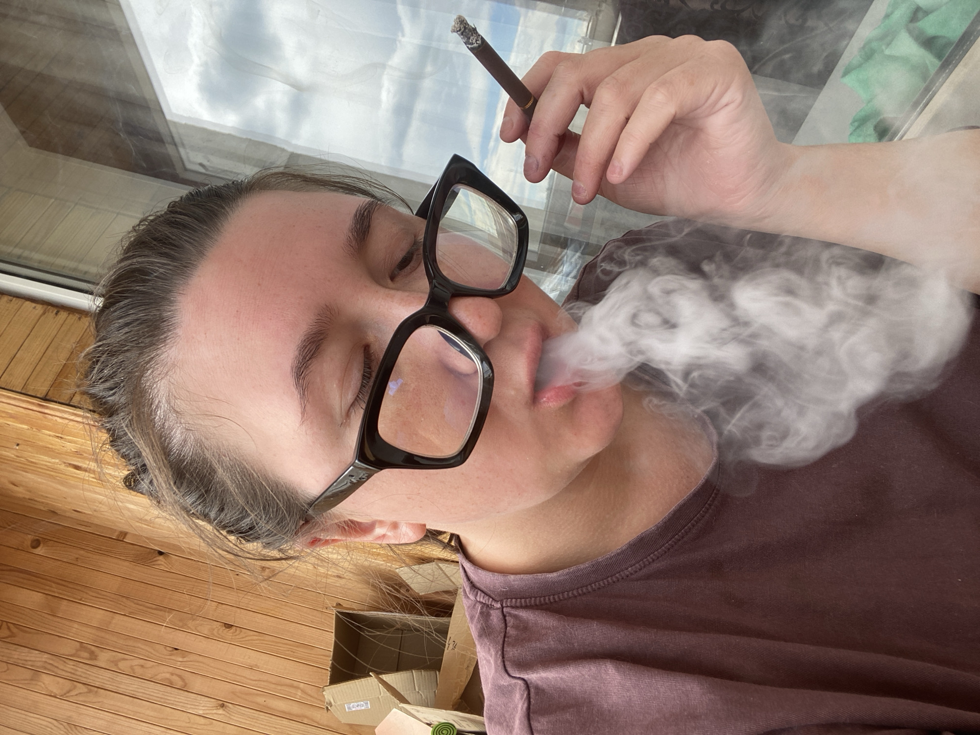The image shows a close-up of a person wearing glasses and smoking a cigar. The person is exhaling smoke, which is billowing around their face. They are wearing a purple t-shirt. The background appears to be a wooden wall and a window with clouds visible outside. The scene suggests an indoor or semi-outdoor setting, possibly a balcony, on a cloudy day. There are cardboard boxes visible in the background, suggesting a storage area or a location where items are being unpacked. The overall impression is a candid, everyday moment captured on camera. The location of Chaikovsky, Russia is not discernible from the image itself.