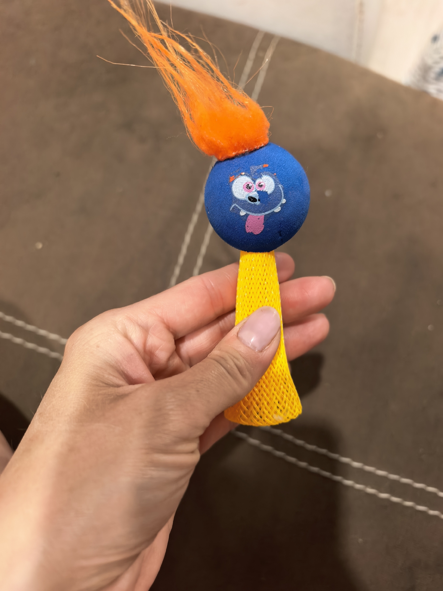 A hand holds a toy. The toy has a blue, round head with a cartoonish face and orange, fuzzy hair. Below the head is a yellow, cone-shaped body with a net-like texture. The background is a brown surface with white stitching. The location is Unknown, Unknown.