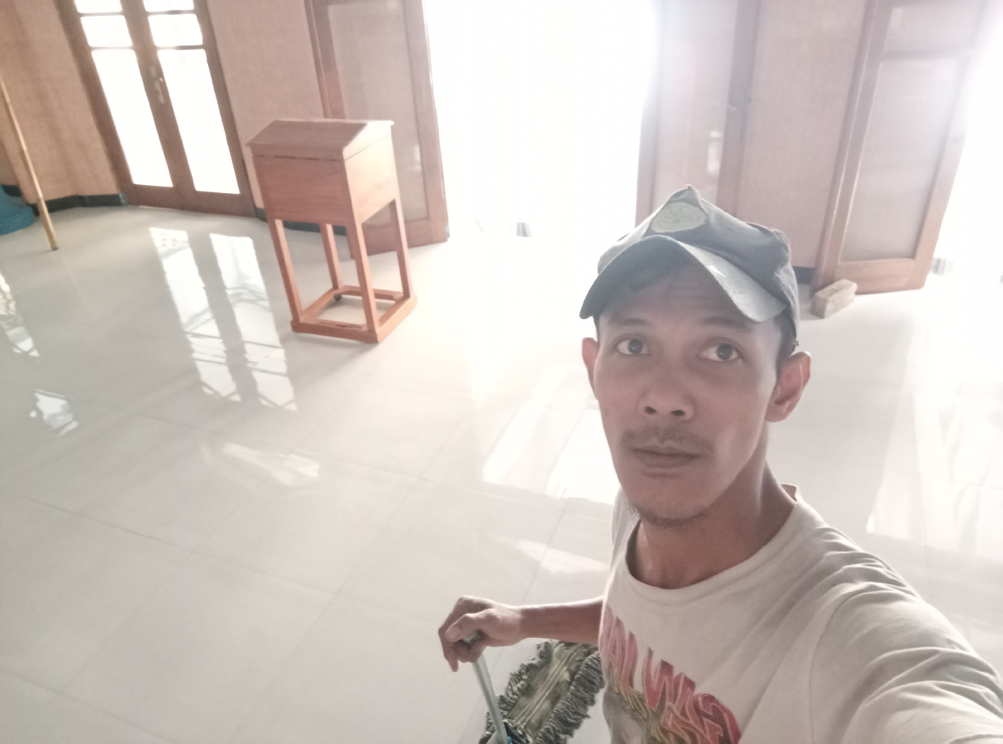 A man wearing a baseball cap and a t-shirt is in a well-lit room, possibly a house or building, in Ngablak, Indonesia. He is holding a mop. The room has a tiled floor that reflects the light. In the background, there are wooden doors and a wooden object with legs. The overall impression is that the man is engaged in cleaning activities within the room.