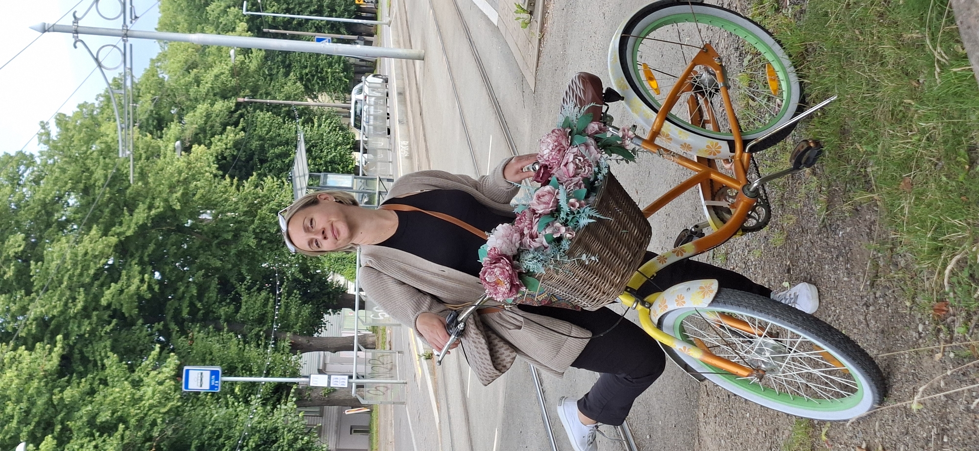 A woman with short blonde hair, wearing a beige cardigan over a black top and black pants, poses with a vintage-style bicycle. The bicycle is orange and yellow with white fenders adorned with floral patterns and has a wicker basket in front filled with pink and white artificial flowers. She is leaning back slightly, with her left leg extended. The scene appears to be in an urban setting in Tallinn, Estonia, indicated by a blue bus stop sign visible in the background, which is a common sight in the city. There are tram tracks on the road and trees lining the street, with a bus shelter in the distance. The ground is a mix of gravel and grass. The lighting suggests an overcast day.
