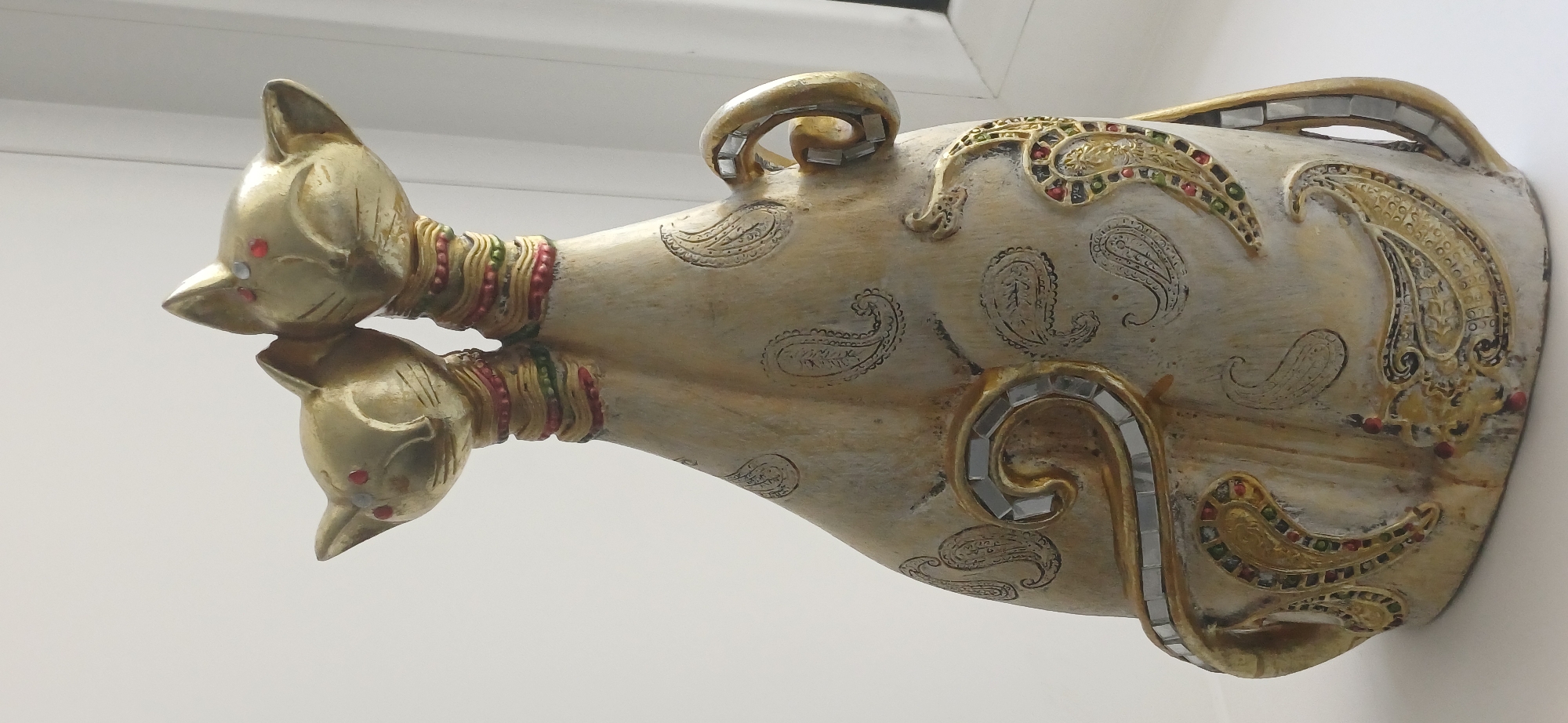 A decorative statue of two golden cats with red eyes sits on a white surface. The cats are facing forward and are positioned close together, with their bodies forming a single, elongated shape. The statue is decorated with paisley patterns in black and gold, and various colorful gemstones are embedded in the gold accents. The background is a plain white wall, with a hint of a window frame on the right side. The lighting suggests the image was taken indoors during the daytime. The statue appears to be placed on a windowsill or a similar flat surface. There is no information available to determine its location in the city of Ekibastuz, Kazakhstan.